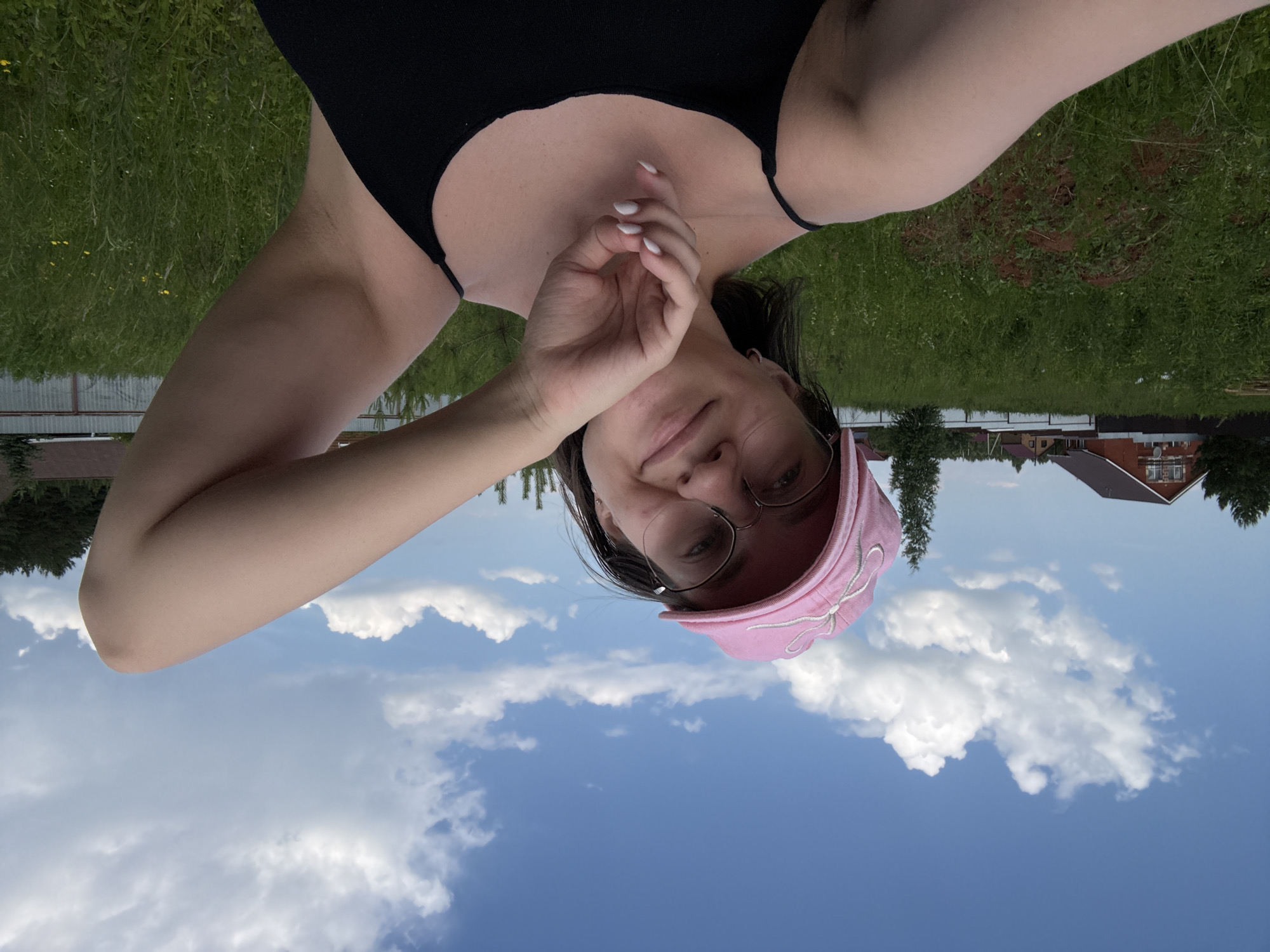 A selfie taken outdoors under a blue sky with white clouds shows a young woman wearing a pink baseball cap and glasses. She is looking towards the camera with a slight smile, her right arm raised and bent at the elbow. She is wearing a black tank top. In the background, there is a grassy field with scattered small yellow flowers, some trees, and houses with reddish-brown roofs. The scene appears to be in a rural or suburban area, possibly on the outskirts of a city or town, and is not specifically identifiable as being in the city of Dokshytsy, Russia based on visual cues alone.
