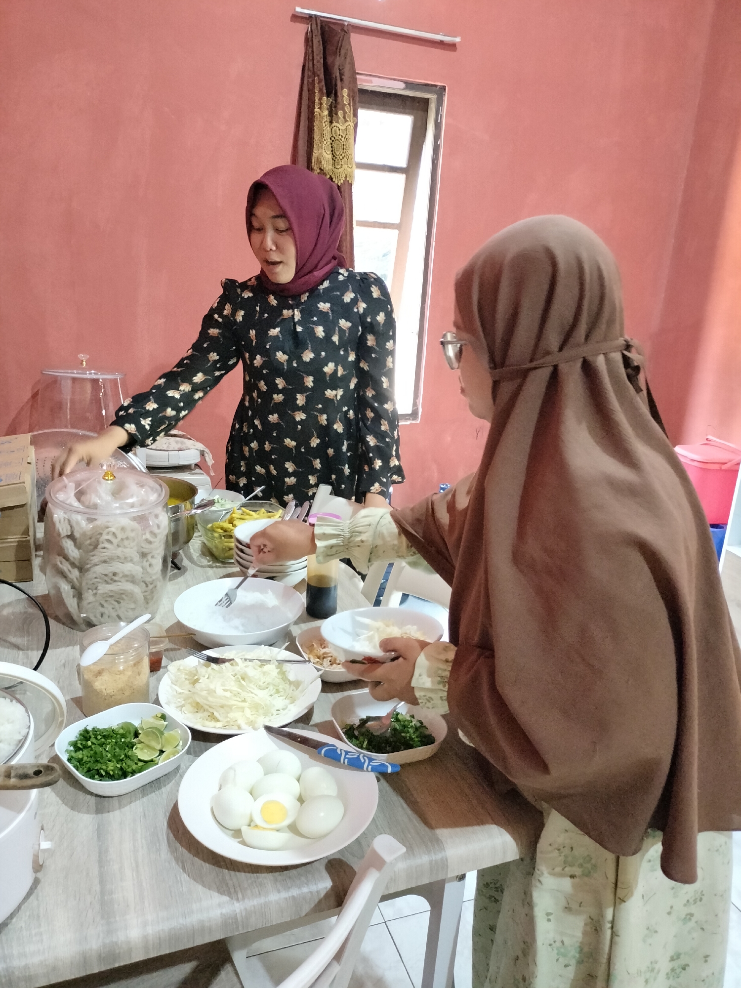 In Angsana, Indonesia, two women prepare food at a table. The woman on the left, wearing a burgundy hijab and a black floral dress, stands behind a table laden with various dishes. She is reaching towards a large glass jar filled with what appear to be crackers. To her right, on the table, are bowls containing chopped green vegetables, sliced limes, shredded cabbage, and boiled eggs. A large container of rice is visible to the left of the jar. The woman on the right, wearing a brown hijab and a light green floral dress, is seated and appears to be serving food from a small plate into a bowl. She is wearing glasses and holding a fork. In front of her is a small bottle of dark sauce and another plate with some food. The table is set in a room with pink walls and a window with brown curtains in the background.