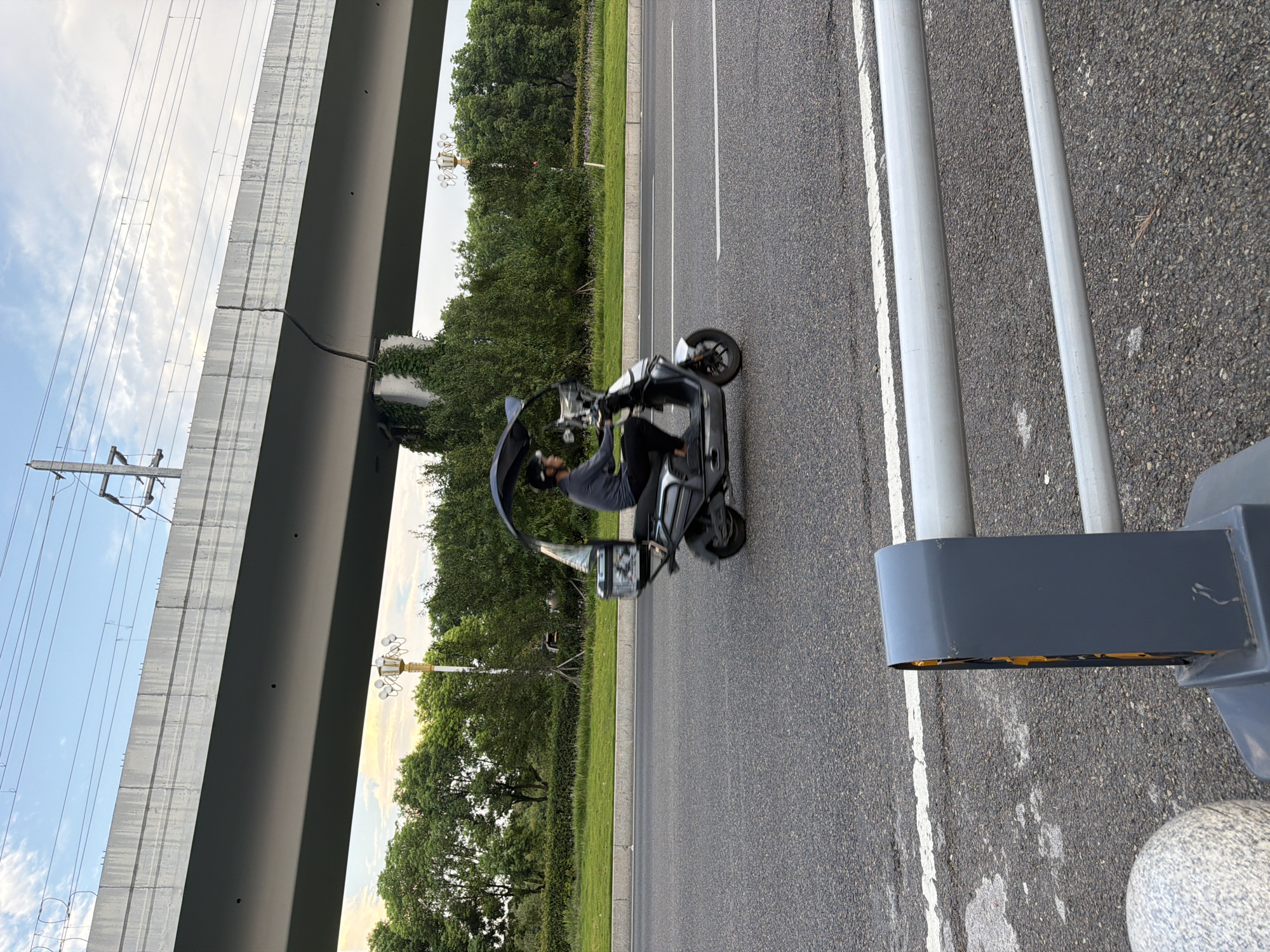 A person is riding a three-wheeled electric scooter with a canopy on a paved road in Jiangjin, China. The scooter is silver and has a black canopy extending over the rider. The rider is wearing a grey long-sleeved shirt, black pants, and a helmet. The scooter has a storage box mounted on the back. In the background, a large concrete overpass runs horizontally across the image. Trees and green grass are visible on either side of the road. Overhead power lines are visible against a cloudy sky. In the foreground, a grey metal guardrail and a grey, textured bollard are partially visible. The scene appears to be from a slightly elevated perspective, looking towards the scooter.