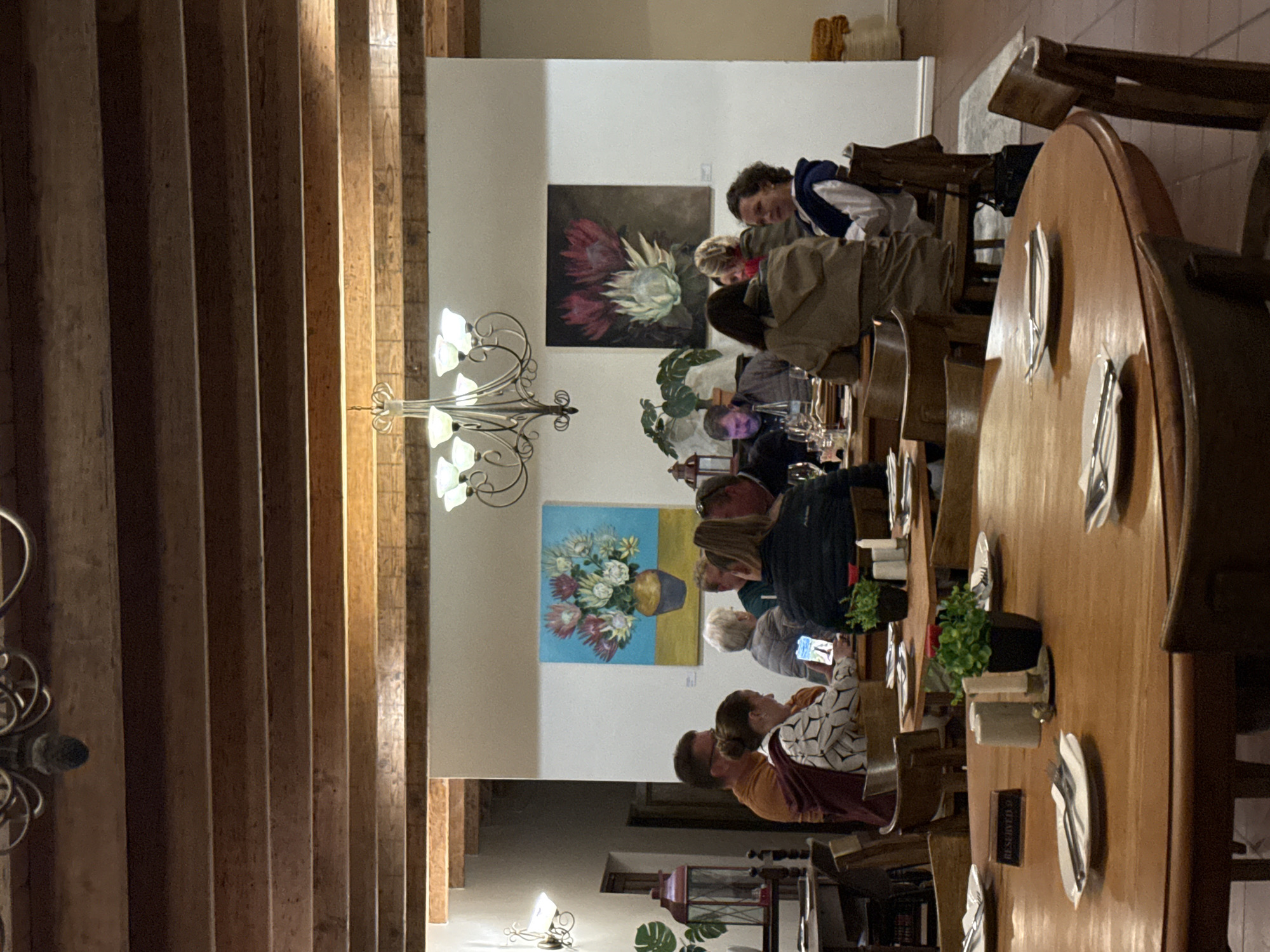 A photograph taken in Paarl, South Africa, shows a long wooden table set for a meal, around which a group of people is seated. The table is in a room with wooden beams on the ceiling and artwork hanging on the walls. The diners are engaged in conversation and other typical dining activities.