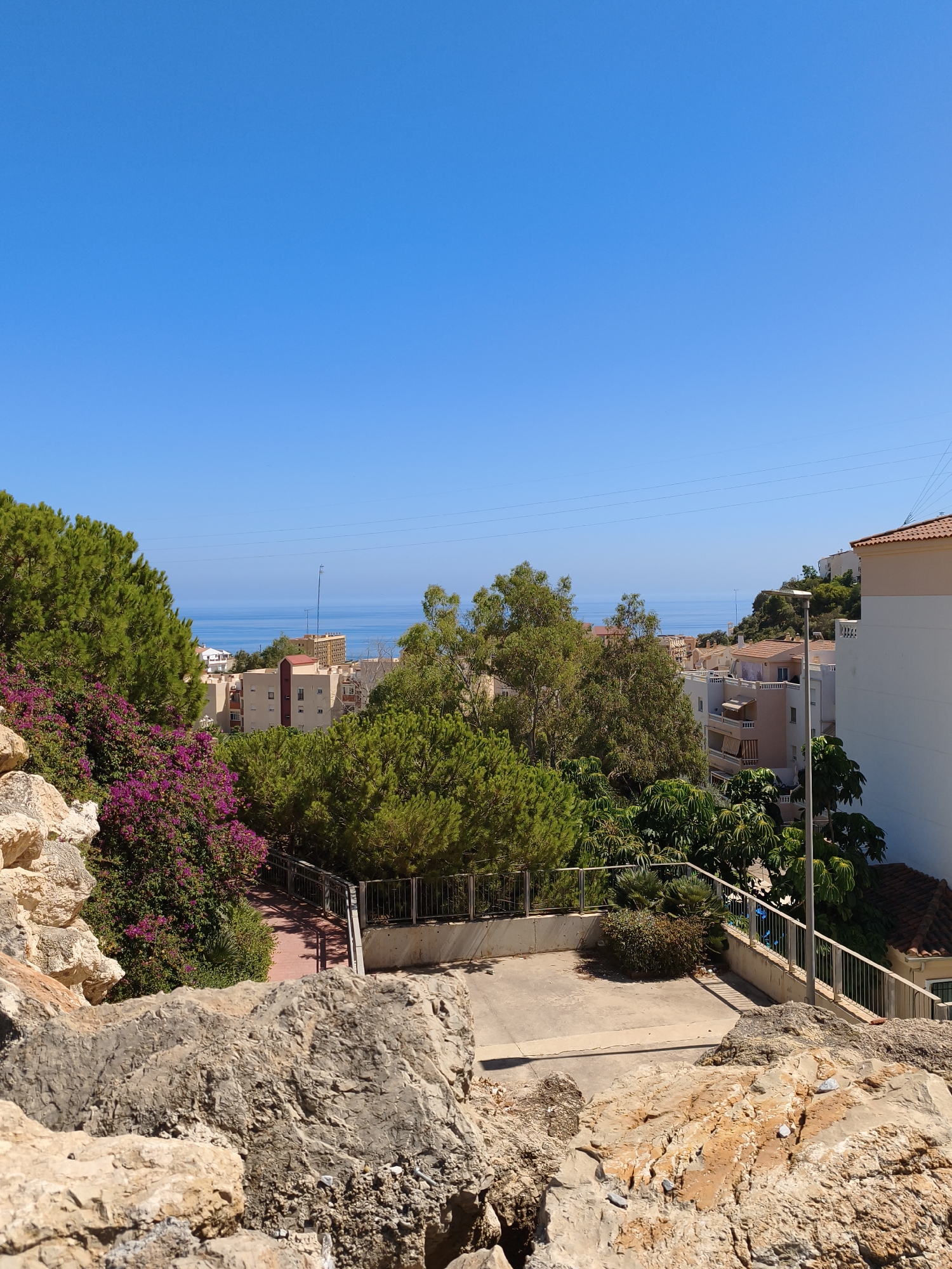 This is a photograph taken from a rocky, elevated viewpoint overlooking a coastal town. The sky is a clear, bright blue, extending to the horizon where it meets the calm, deep blue sea. In the middle ground, a cluster of multi-story buildings with cream-colored facades and terracotta roofs are nestled amongst lush green trees and vibrant pink bougainvillea. Some buildings have balconies and awnings. A paved pathway with a metal railing winds down the slope in the foreground. Further into the distance, more buildings and greenery are visible, with the sea stretching out beyond them. The foreground is dominated by large, rough-textured rocks, some lighter and sandy in color, others darker and gray. The overall impression is a sunny, Mediterranean landscape.