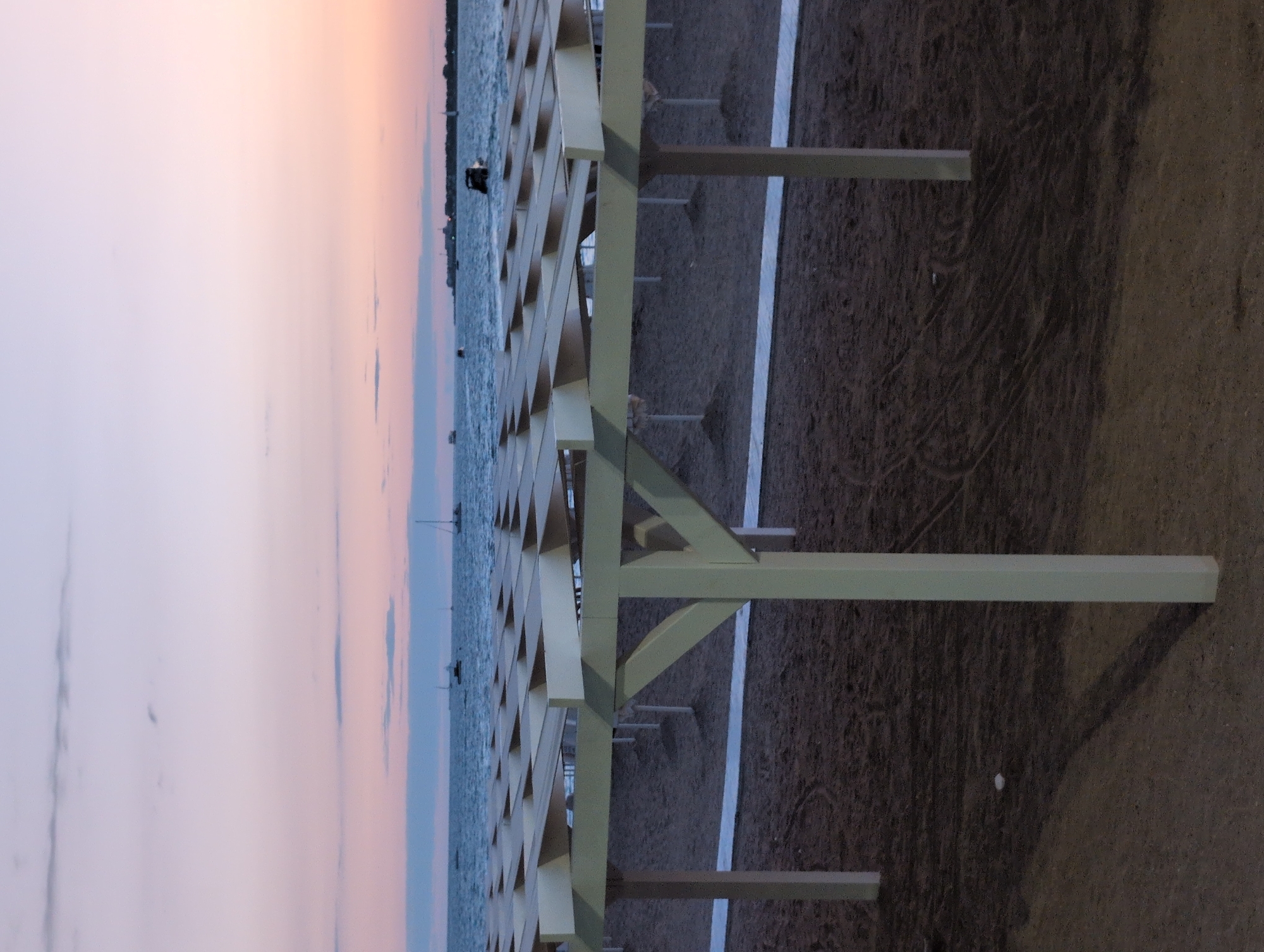 A beach with rows of empty sun shelters stands under a pastel sky at dusk, with the calm sea stretching to the horizon. The sand is dark, marked with faint tire tracks from beach maintenance vehicles. A white mat or walkway runs along the front of the sun shelters. In the distance, several sailboats and a motorboat are visible on the water, and a faint line of buildings suggests a distant shore. The scene appears to be the waterfront in Gelendzhik, Russia.