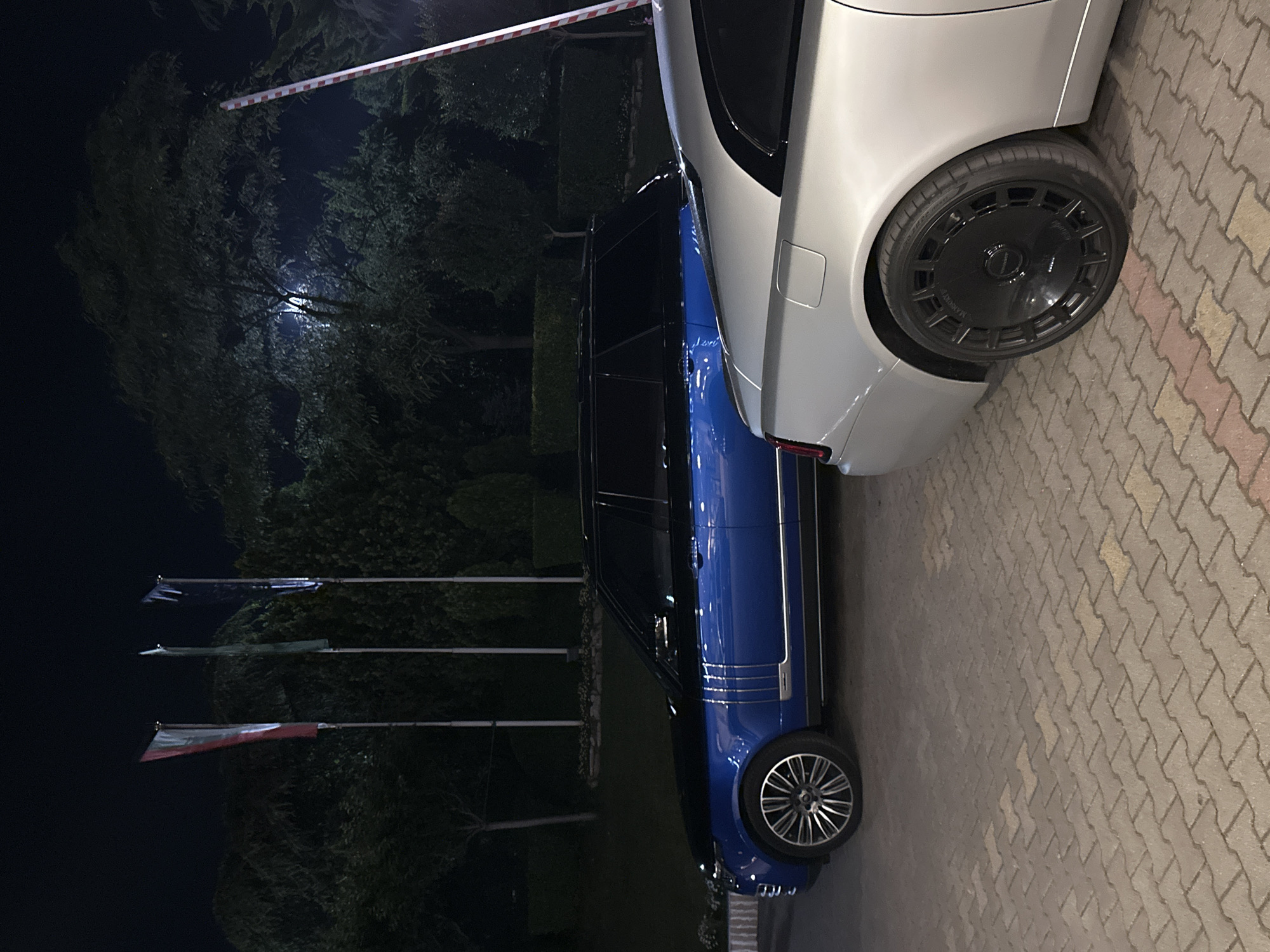 The image depicts a nighttime outdoor scene in Stara Zagora, Bulgaria, featuring two luxury vehicles parked on an interlocking concrete paver surface. On the left, a two-tone blue and black Range Rover is visible, displaying multi-spoke silver and black alloy wheels and chrome vertical accents on its front fender. To the right, partially obscured, is a silver or light grey Rolls-Royce Wraith coupe, identifiable by its distinctive rear quarter panel and a prominent black aftermarket wheel with "MANSORY" branding on its center cap.
In the background, dense, dark green trees and shrubs form a landscaped environment. Three flagpoles stand erect in the left-center, bearing flags that are indistinct due to the low light, though the leftmost flag appears to have red and white elements. A red and white striped automatic barrier arm is partially visible extending from the upper right corner. A bright, diffused light source is discernible within the foliage in the upper-middle section of the image. The ground is paved with brown and grey interlocking concrete blocks. No individuals or specific activities are present in the scene.