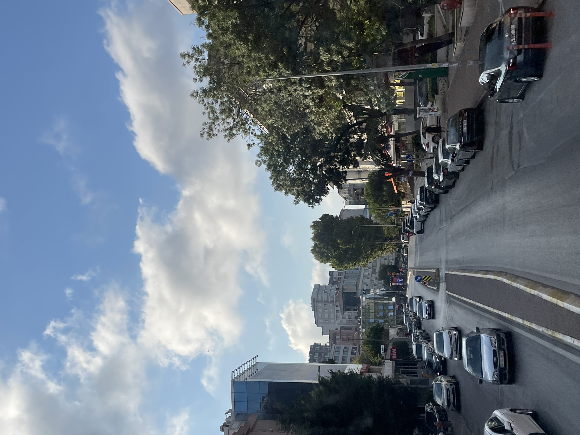 The image depicts a view down a city street in Kadıköy, Turkey, on a partly cloudy day. The street is lined with buildings of various architectural styles, including modern glass-fronted structures and older, multi-story apartment buildings. Lush green trees are interspersed throughout the urban landscape, casting shadows on the pavement. Numerous cars are present on the road, some driving and others parked along the sides, indicating moderate traffic. Pedestrians are visible on the sidewalks, and streetlights line the thoroughfare. The sky is a mix of blue and white clouds, suggesting pleasant weather. A sign for "BİM," a Turkish supermarket chain, is visible on one of the buildings.