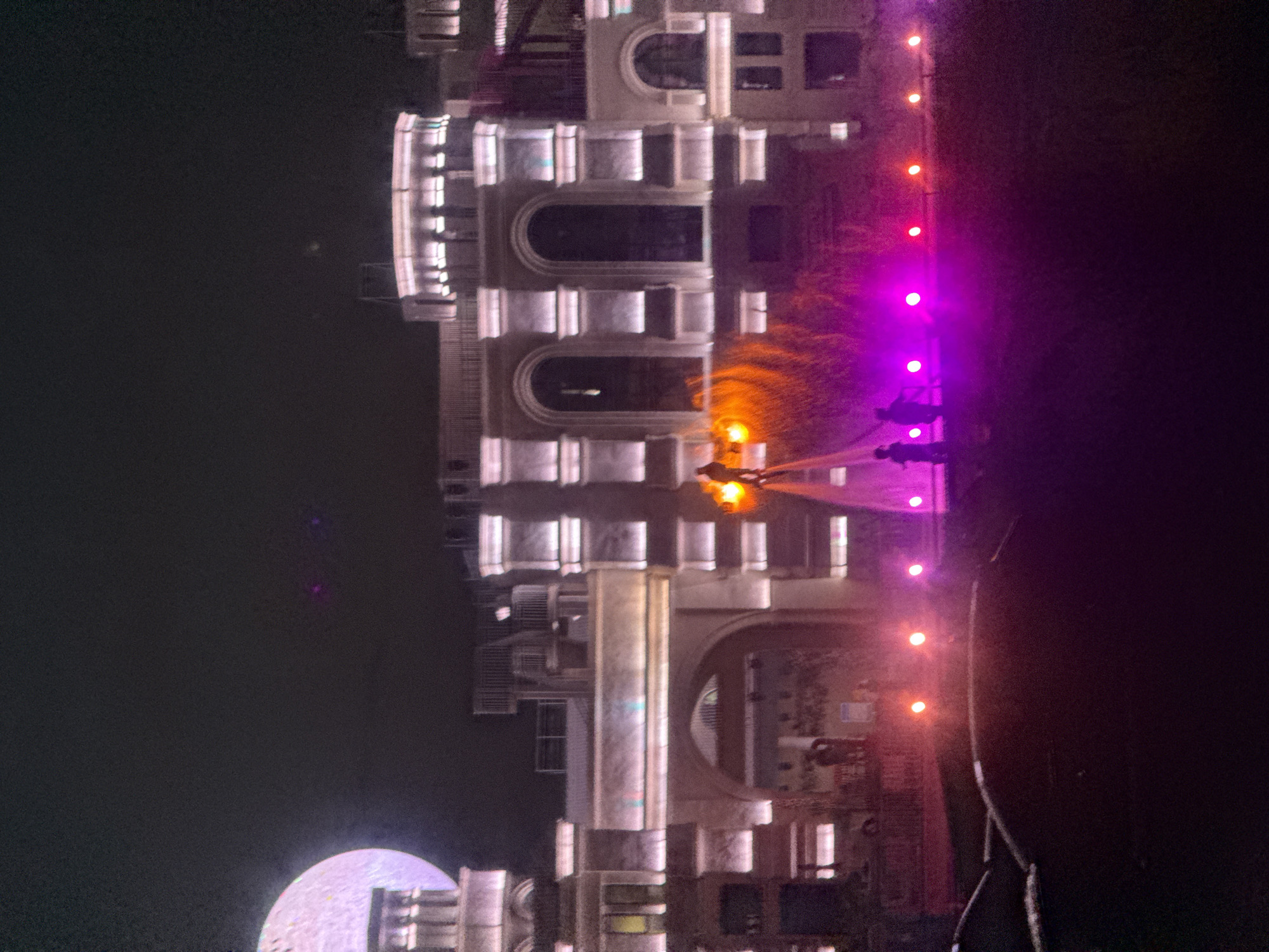 The image depicts an outdoor nighttime scene in Qinhuangdao, China, featuring a large, illuminated building with architectural elements resembling a castle or palace. The building is lit with bright white and pink lights, highlighting arched windows and decorative facades.
In the foreground, there is a large, dark, amorphous object that appears to be the hood of a car. Scattered across the ground in front of the building are various colored lights, predominantly pink and red, creating a vibrant, atmospheric effect.
In the middle ground, a water-jet performance is underway. Two figures are silhouetted against a backdrop of orange and pink light, appearing to be propelled by water streams. One figure is positioned higher, seemingly standing on the water jets.
To the left of the building, a large, round, illuminated object, resembling a stylized moon or a giant orb, is visible, casting a pinkish glow.
Several indistinct figures are visible in the background, near the base of the building and around the water performance area, suggesting an audience or participants in the event. The overall scene conveys a festive or entertainment-oriented atmosphere.