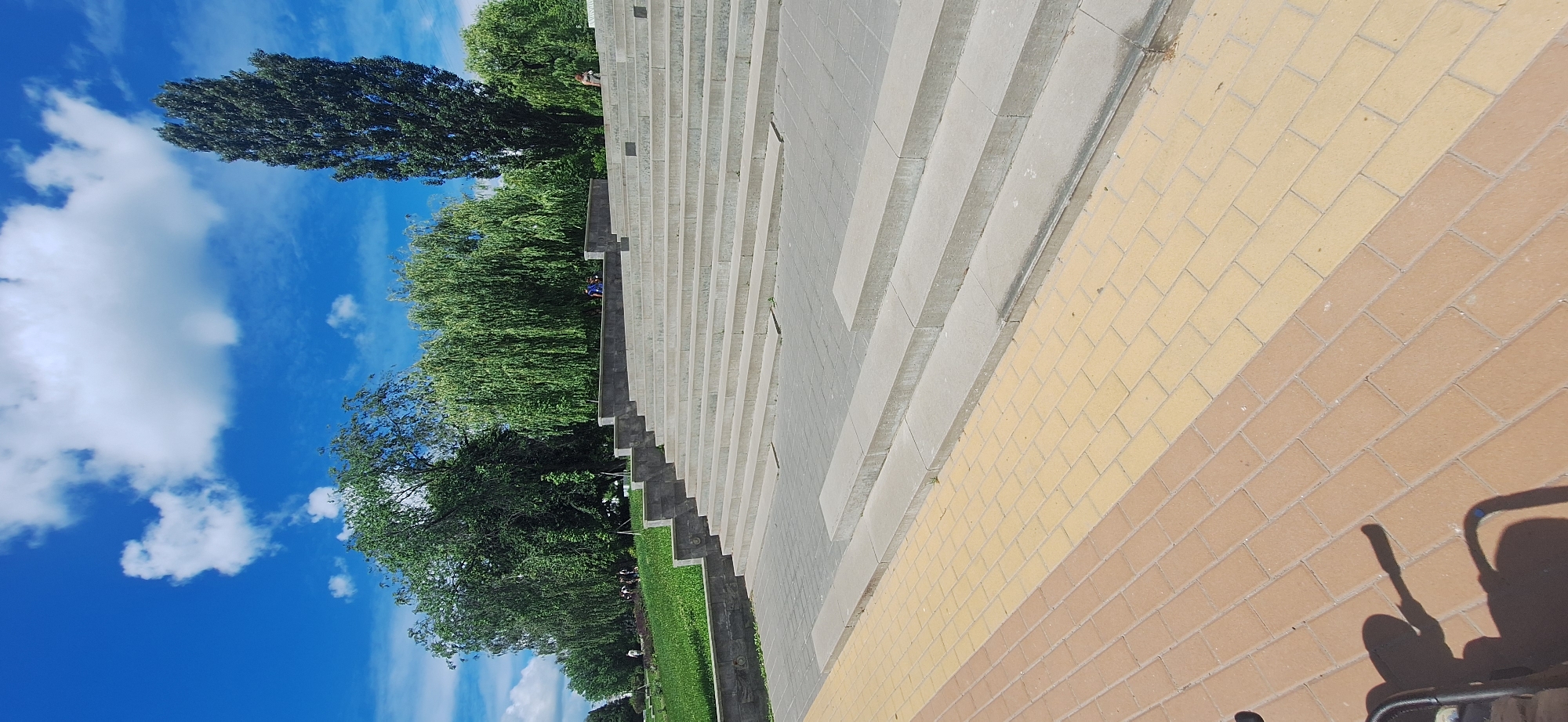 A wide, outdoor, ground-level shot shows a large set of stone stairs with people scattered across them and in the distance. The stairs are concrete and are lined by a walkway paved with tan and yellow bricks. The sky is bright blue with several fluffy white clouds, and lush green trees with weeping branches and one tall, slender tree frame the background. To the left of the stairs, a green lawn slopes downwards and is bordered by a stone wall. The image appears to have been taken on a sunny day, possibly in the late spring or summer. This scene is likely in Bila Tserkva, Ukraine, given the description of the location.