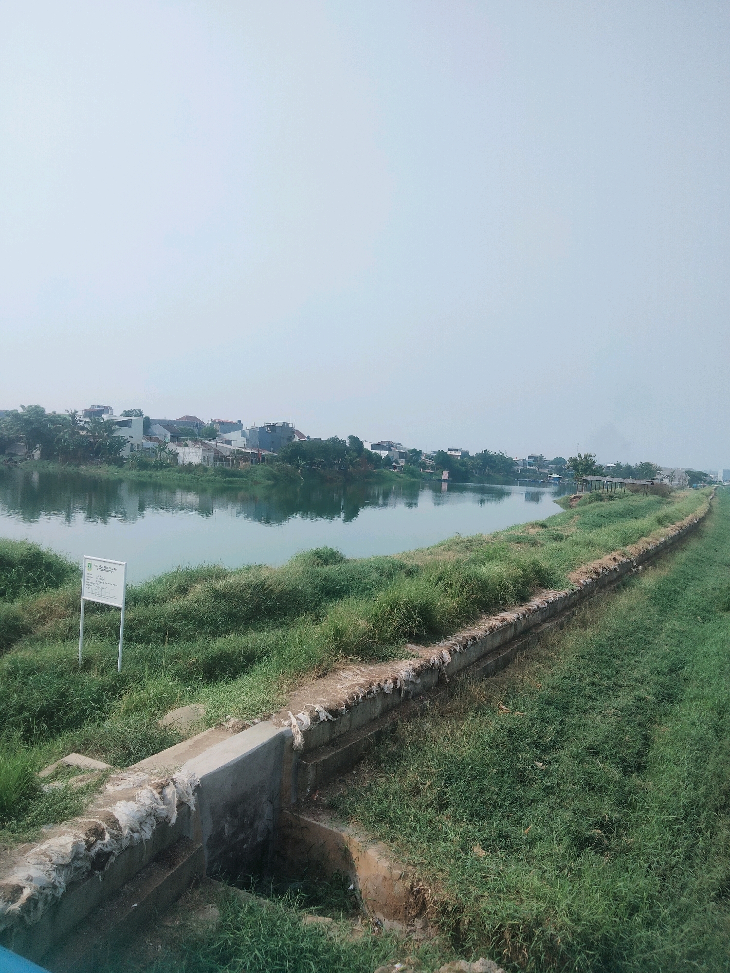 The image depicts a body of water, possibly a river or lake, with a grassy embankment along its edge. In the background, a dense line of buildings, including houses and other structures, is visible, suggesting an urban or suburban setting. The sky is overcast and a light blue-gray. In the foreground, there is a concrete structure with what appears to be some sort of drainage or water management system. A white sign with some text and graphics is positioned in the grass on the left side of the image. The overall scene suggests a somewhat developed area adjacent to a natural water feature. The location is identified as being in the city of Tangerang, Indonesia, a significant industrial and commercial hub on the island of Java.