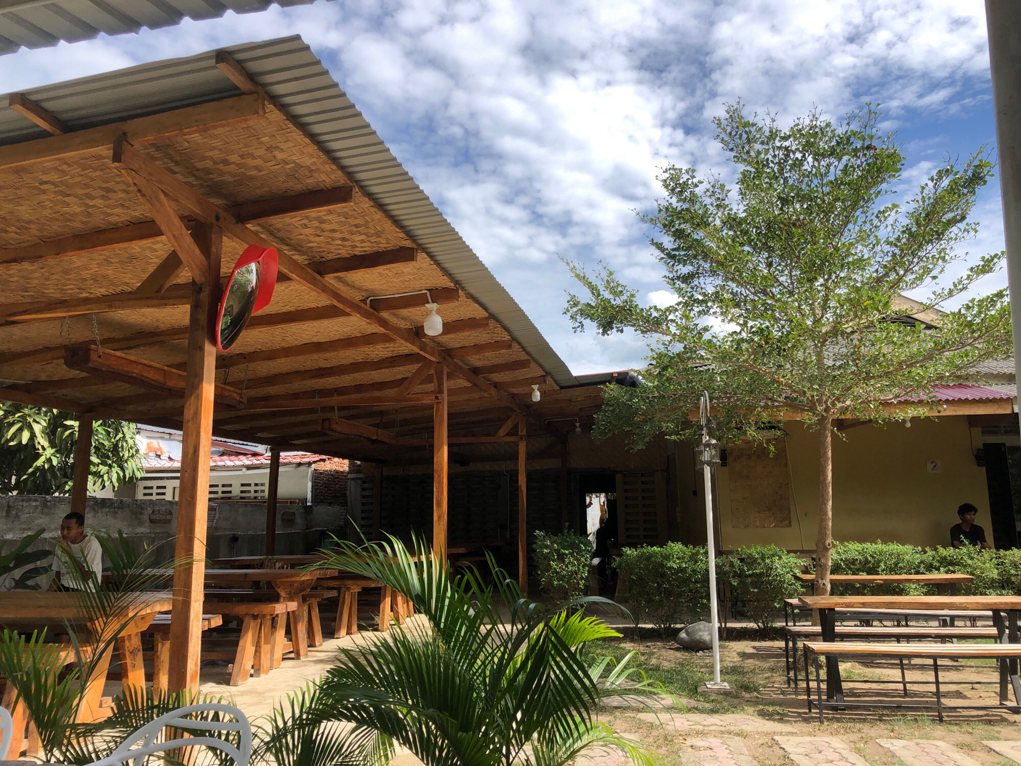 The image depicts an outdoor seating area at a restaurant in Tambonbaroh, Indonesia. The area is covered by a rustic, sloped roof constructed from corrugated metal and bamboo thatch, supported by wooden beams. Several long wooden tables with benches are arranged on a sandy ground, with some tables closer to the foreground and others further back. A few individuals are visible; one person in a light-colored shirt is seated at a table on the left side of the frame, and another person in a dark shirt is seen sitting on a bench near a building on the right. Green foliage, including palm fronds in the foreground and a medium-sized tree on the right, adds to the natural environment. The background shows parts of buildings, including one with a visible house number "2". The sky is partly cloudy, with white clouds against a blue backdrop. A red convex mirror is mounted on a wooden post under the covered area. The overall scene suggests a casual dining or gathering space in a tropical setting.