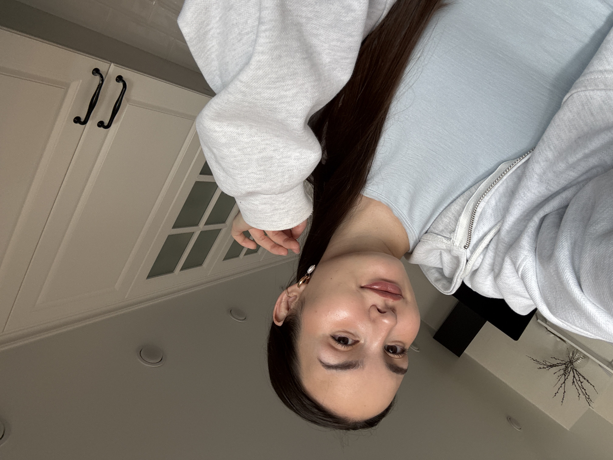 A close-up indoor shot features a young woman posing for a photo, depicted upside down due to a 90-degree counter-clockwise rotation of the image. She has long, dark hair and is wearing a light blue top underneath a light grey zippered hoodie. Her right ear, adorned with a gold hoop earring featuring a pearl drop, is visible. She has a clear complexion, defined eyebrows, and a subtle, pleasant expression as she looks directly at the camera.
The background shows elements of a home interior, likely in Chaikovsky, Russia. White kitchen or utility cabinets with dark, ornate handles are visible behind her. Above the cabinets, parts of a ceiling with recessed lighting and cabinet doors with a grid-patterned glass design can be seen. To her right, a decorative arrangement of thin, dark branches in a vase sits on a surface, next to a dark, rectangular object. The lighting is soft, suggesting an indoor setting. There is no visible text in the image.
