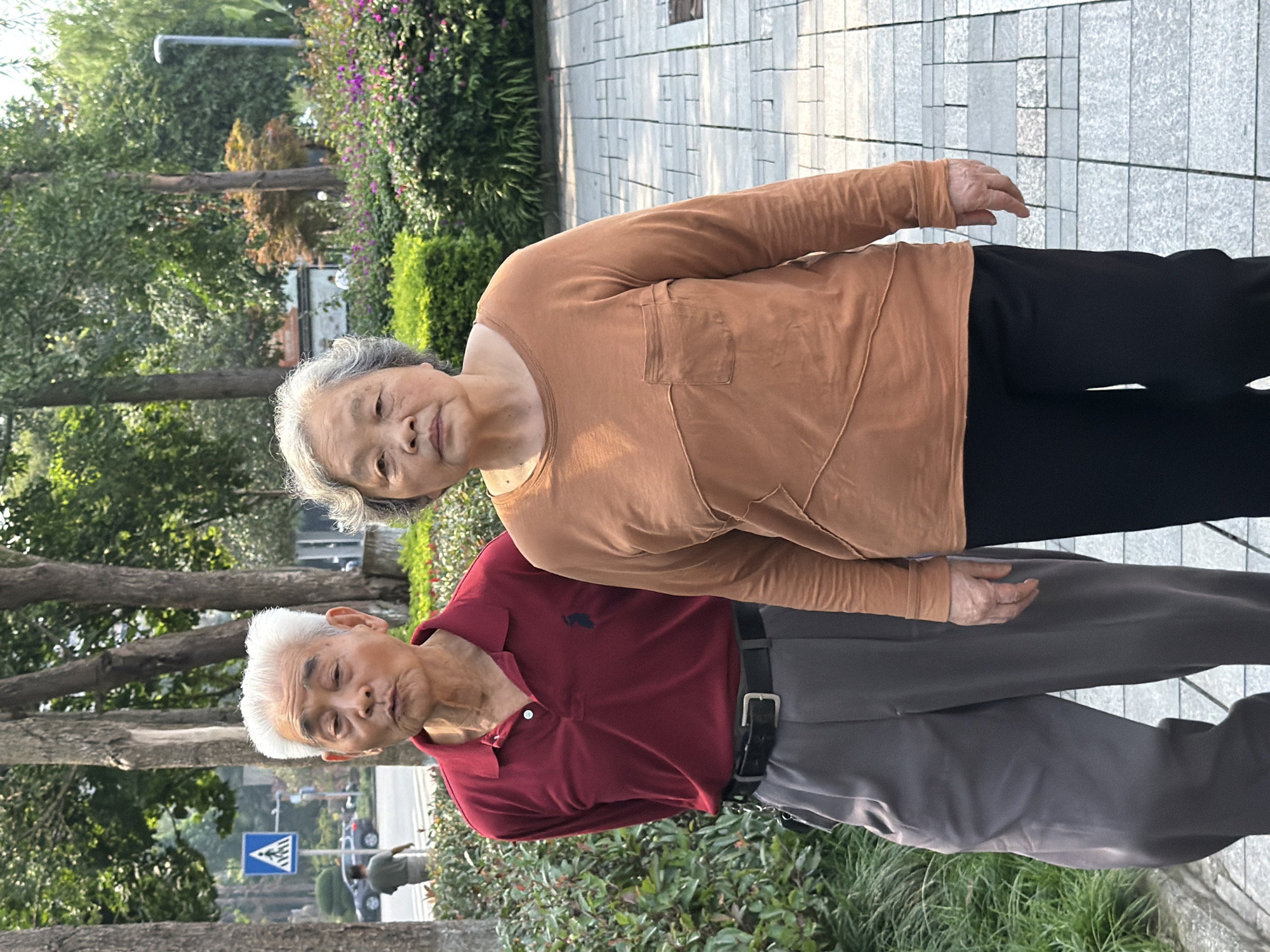 The image shows an elderly Asian couple walking side-by-side on a paved sidewalk in what appears to be a park or a tree-lined street in Jiangjin, China. The man is on the left, wearing a maroon polo shirt, grey trousers, and a black belt. He has white hair and is looking towards the camera with his mouth slightly open. The woman is on the right, wearing a long-sleeved rust-colored top with a chest pocket and black trousers. She has grey hair and is looking forward with a neutral expression. The background features lush green trees and bushes, with some flowering plants in the mid-ground. A street sign with a pedestrian crossing symbol is visible in the distance to the left, along with a car and a person walking on the road. The paving stones of the sidewalk are a light grey color with some darker square accents. The lighting suggests it is daytime, possibly late morning or early afternoon.