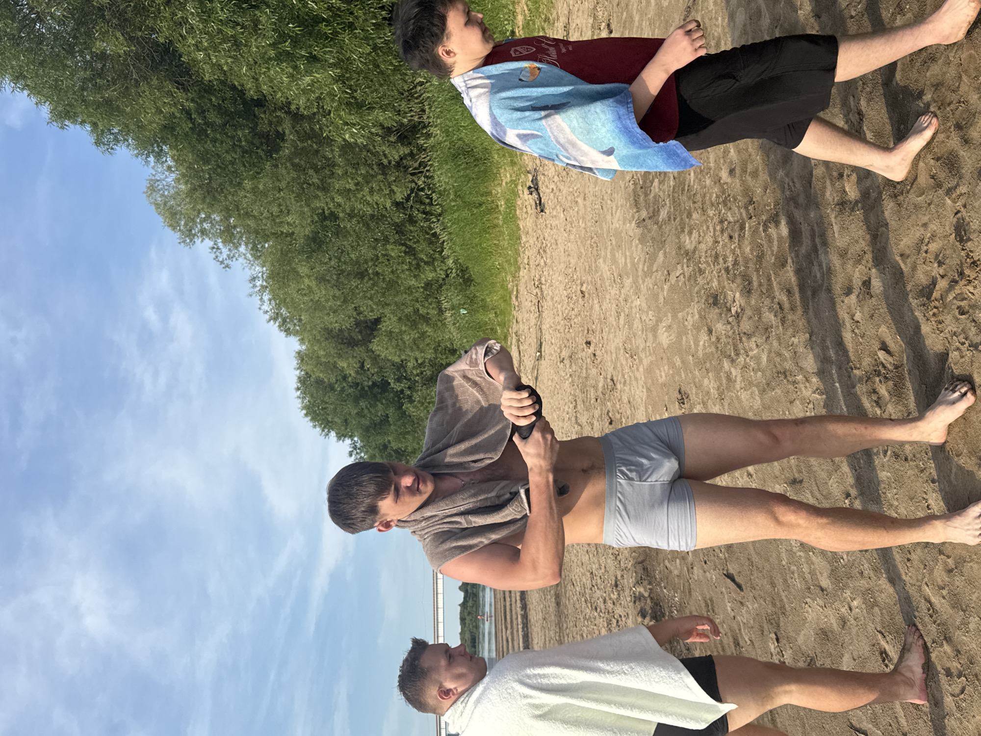 Three young men are standing on a sandy beach. The man in the center is wearing grey boxer briefs and has a tan towel draped over his shoulders. He is holding a black object in his hands, possibly a phone or a small device, and appears to be interacting with it. To his left, another man is wearing a white towel around himself and dark swim shorts. He is looking towards the man in the center. To the right, a third man is walking away from the camera, wearing a maroon t-shirt, black shorts, and a blue towel with a dolphin pattern draped over his shoulders. The background shows a body of water with a bridge and a small boat in the distance, and lush green trees line the edge of the beach. The sky is blue with scattered clouds. The location is the beach in Ryabinino, Russia.