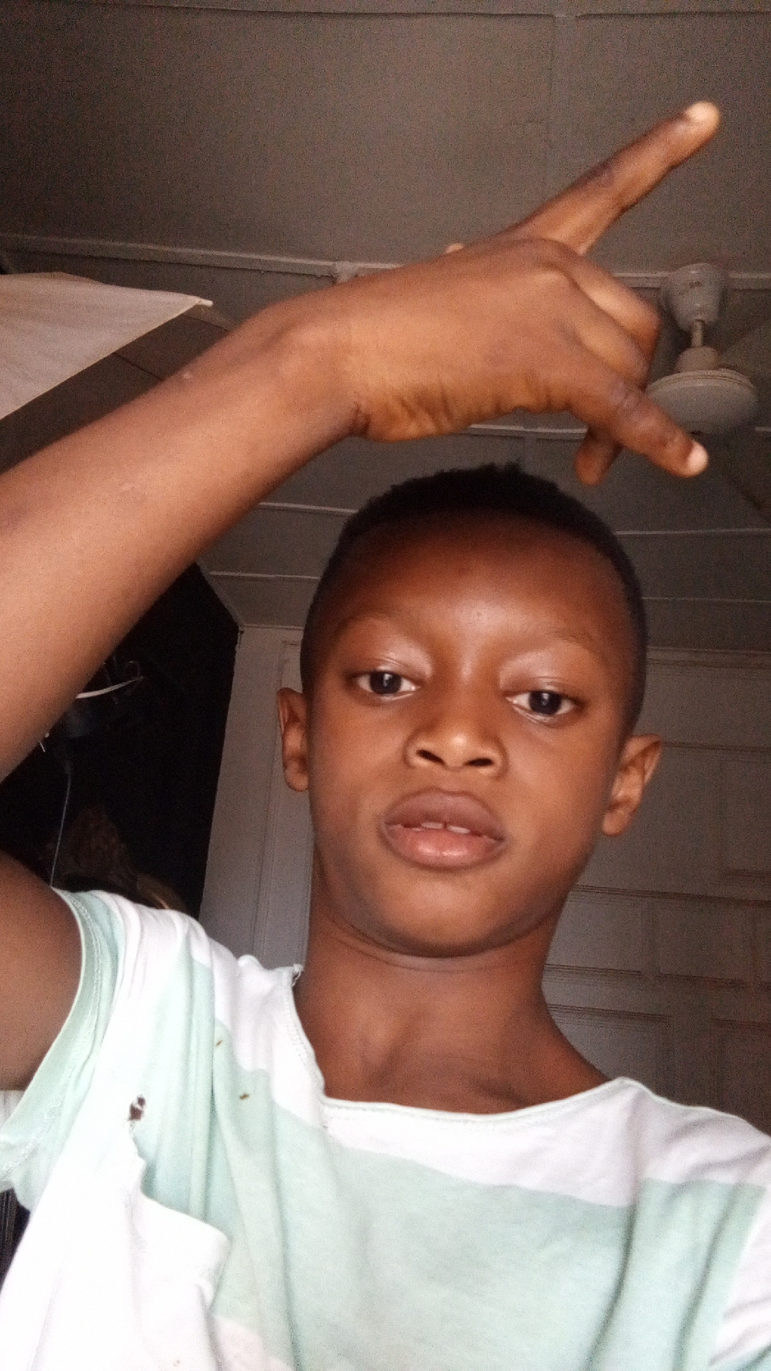 A portrait-style photograph captures a young person, likely male, with dark skin and closely cropped hair, looking directly at the camera with a neutral expression. His face is central in the frame, and his mouth is slightly open.
He is wearing a t-shirt with horizontal stripes, alternating between white and a light mint green or pale teal color. His left arm is raised above his head, with his hand making a gesture where the index finger and pinky finger are extended upwards, while the middle and ring fingers are curled down towards the palm.
The setting is indoors, with a wall visible in the background, appearing to be a light grey or beige color, with a white trim line. A white ceiling fan is partially visible in the upper right corner of the frame. To the left of the subject, a darker area suggests an open doorway or a dark part of the room. The lighting appears to be artificial, illuminating the subject's face while the background remains somewhat dim.
The image portrays a casual, posed moment. There is no specific activity or event other than posing for the camera. The time of day cannot be determined from the indoor lighting. The location context indicates this scene takes place in Sabon Tasha, Nigeria. No text is visible in the image.