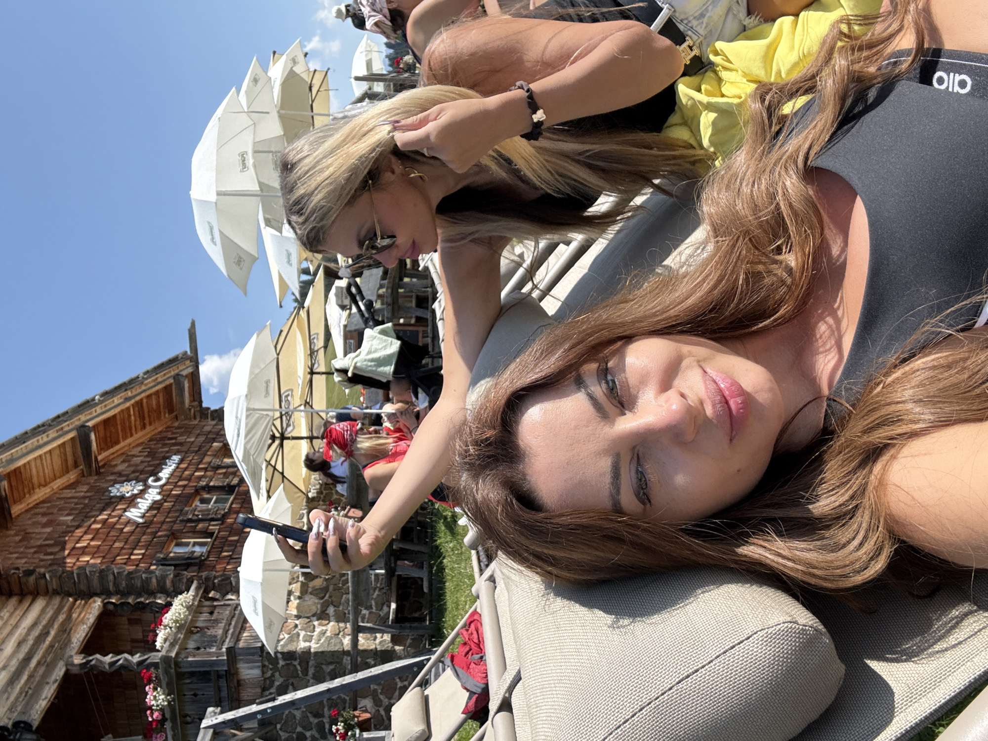 Two women are captured in a selfie taken outdoors, likely at "Malga Confin" in Kastelruth - Castelrotto, Italy, as indicated by signage on the wooden building in the background. The woman in the foreground, with long, wavy brown hair, is reclining on a lounge chair and holding a smartphone to take the photo. She is wearing a black tank top. Beside her, slightly out of focus, is another woman with long blonde hair, wearing sunglasses and a black top, and she appears to be adjusting her jewelry. The scene is set on a sunny day, with white umbrellas providing shade in the background. The environment includes a rustic wooden building with stone accents, a grassy area, and what appears to be outdoor seating.