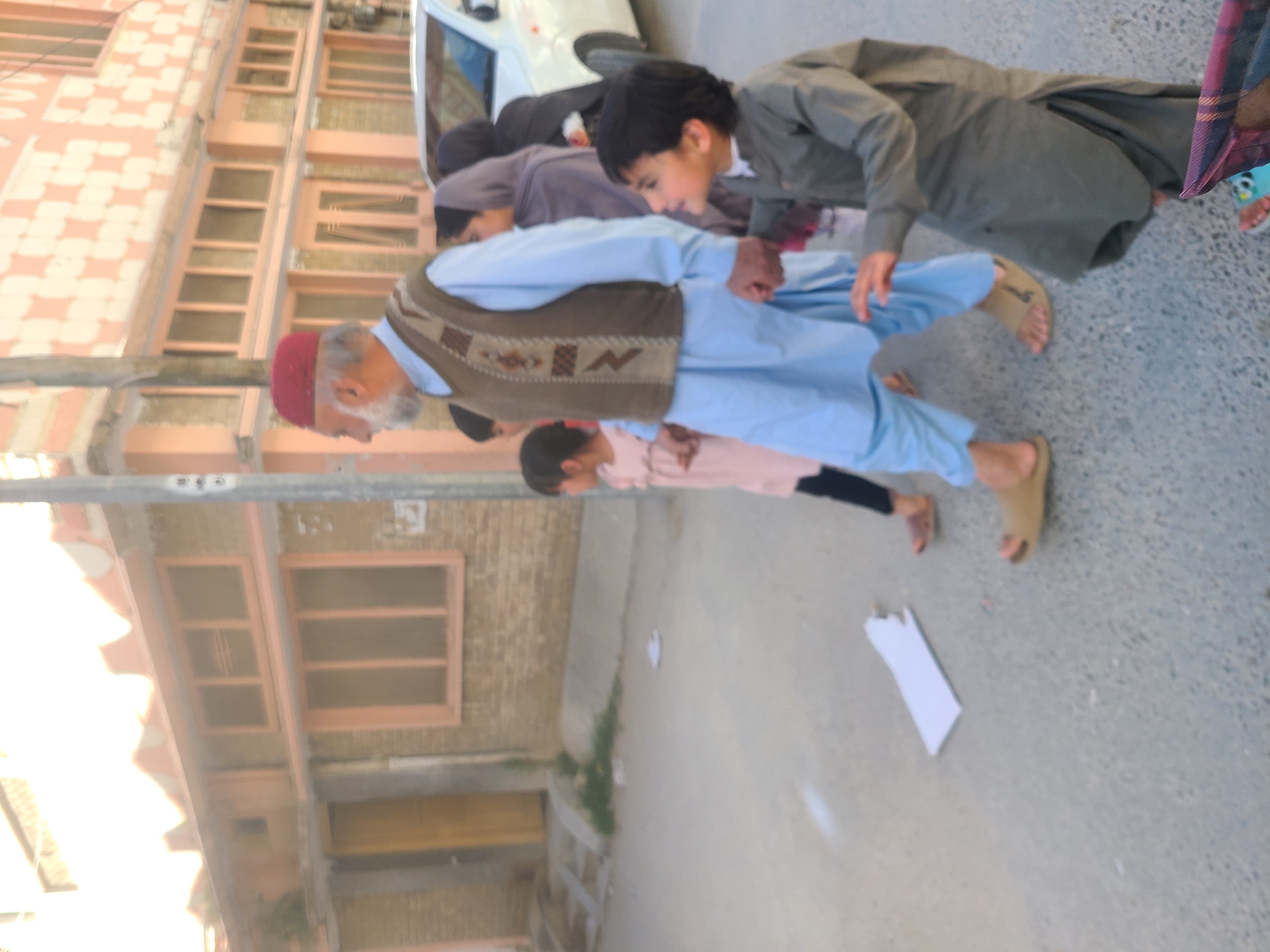 Several individuals are engaged in a physical activity, possibly a game, in what appears to be a street or courtyard. They are arranged in a line, with each person holding onto the person in front of them. The location shows a building with window frames and other urban fixtures, suggesting an urban environment. A white piece of paper sits on the ground. The image was taken in Quetta City Tehsil, Pakistan.