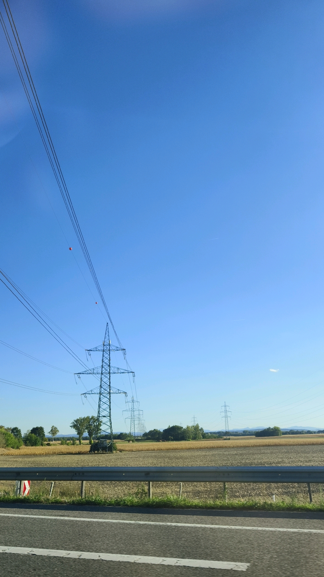 The image shows a clear blue sky over a rural landscape with a series of electricity pylons stretching into the distance. The pylons are tall, lattice-metal structures supporting high-voltage power lines. Two orange spherical markers are visible, attached to the power lines for aviation safety. In the foreground, a road with a white line marking is visible, with a metal guardrail separating it from a field. The field appears to be agricultural, with dry crops. Trees and a small cluster of buildings are visible in the middle ground. The location is identified as Neustift im Felde, Austria. The scene is static and depicts a typical rural infrastructure setting on a sunny day.