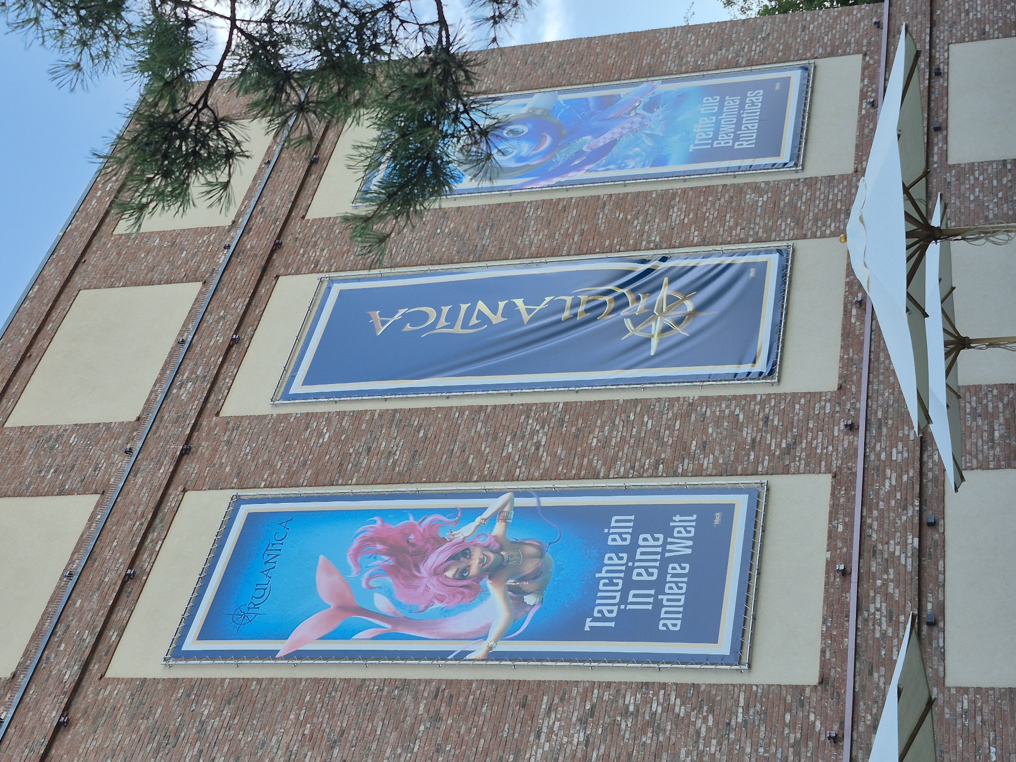 This is a medium-angle, outdoor, eye-level shot of the exterior brick wall of a building in Rust, Germany, likely part of an entertainment venue. Three large vertical banners are displayed on the wall, each framed by a lighter-colored rectangular section. The banners feature animated characters and text.
The leftmost banner displays a cartoon mermaid with pink hair and a pink tail, swimming in a blue underwater scene. Text in German reads: "RULANTICA" at the top and "Tauche ein in eine andere Welt" below the mermaid, translating to "Dive into another world." A small "Mack" logo is present at the bottom.
The middle banner is a solid dark blue with a golden, stylized compass rose logo at the bottom. The word "RULANTICA" is written vertically in gold lettering down the center.
The rightmost banner shows a cartoon octopus-like character with blue skin and large eyes, also in an underwater scene with colorful elements. German text at the bottom reads: "Treffe die Bewohner Rulanticas," translating to "Meet the inhabitants of Rulantica."
The brickwork of the building is visible between the banner sections. The sky is blue with some clouds. A portion of a pine tree's branches is in the upper right corner of the frame, obscuring some of the sky and the rightmost banner. In the foreground, the tops of two large, white, outdoor umbrellas are visible, suggesting an outdoor seating area. The overall impression is of an advertisement for an attraction.