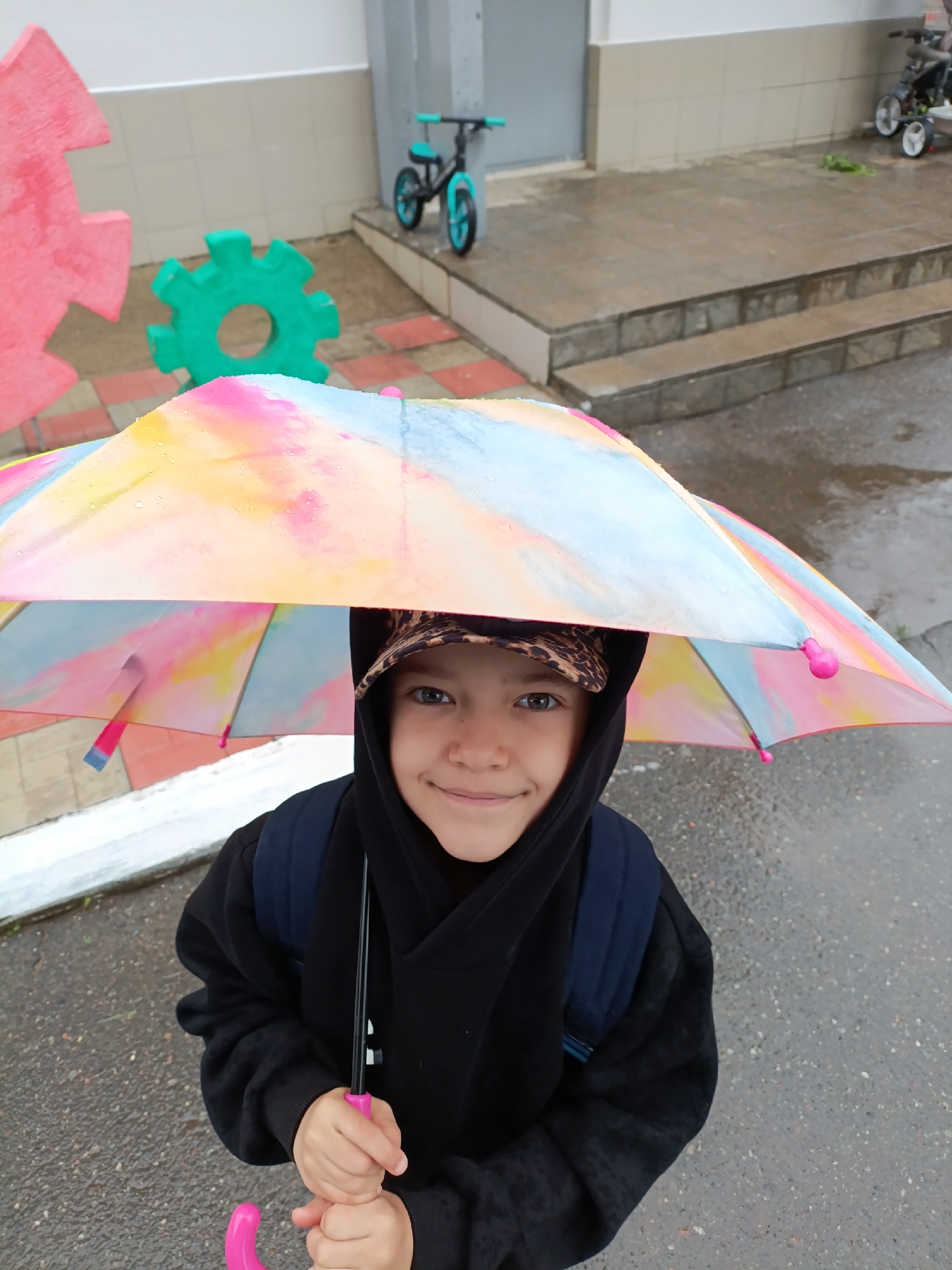 A young person stands outdoors under a colorful umbrella, holding the pink handle. The person is wearing a black hooded sweatshirt, a backpack, and a patterned hat. The ground appears wet, suggesting it is raining or has recently rained. In the background are building walls and a small balance bike. To the left, there are pink and green cog-shaped decorations. The location may be a playground or schoolyard in the city of Chaikovsky, Russia.
