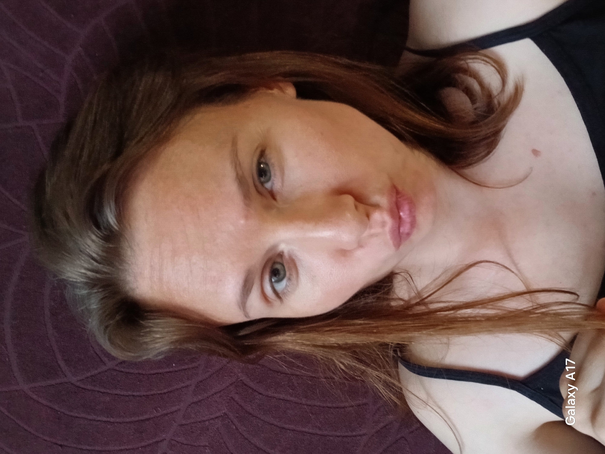 A woman with brown hair lies on a dark purple textured surface. She is wearing a black tank top. Her eyes are light-colored. A mole is visible on her upper chest. The text "Galaxy A17" is present in the lower right corner. There is no information to suggest that the image was taken in Yekaterinburg, Russia.