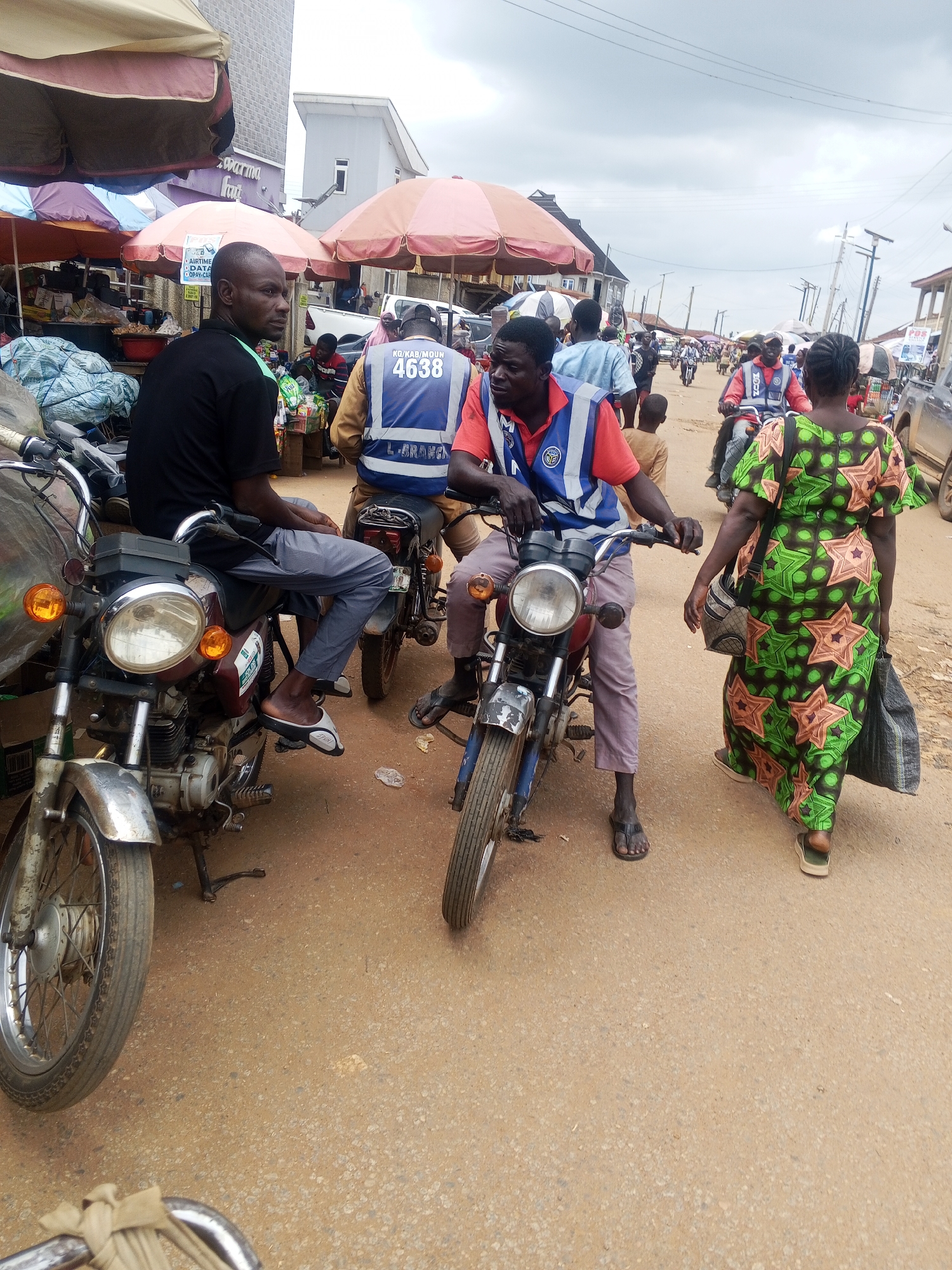 The image depicts a street scene in Kabba, Nigeria, likely a market area given the presence of stalls with umbrellas and merchandise. In the foreground, two men are seated on motorcycles. The man on the left, wearing a black t-shirt and grey trousers, is looking to his right. The man on the right, wearing a red t-shirt with a blue and white vest, is also looking forward. A woman in a green and star-patterned dress is walking away from the viewer towards the right side of the image, carrying a dark bag. Other individuals and motorcycles are visible in the background, suggesting a bustling environment. The ground is a sandy, dirt road, and overhead, the sky is cloudy. The overall impression is of an everyday street scene in a Nigerian town.