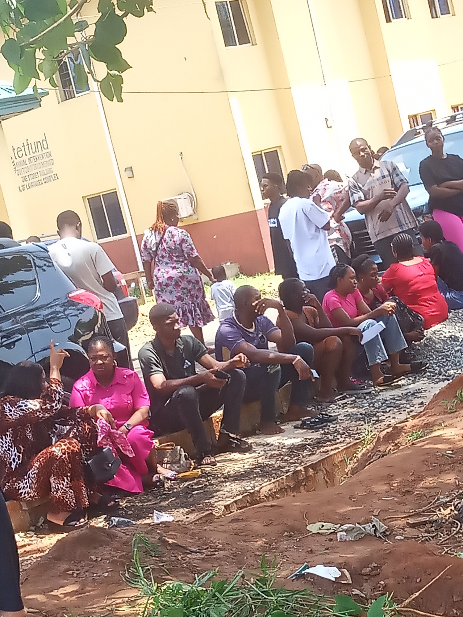 The image shows a group of people gathered outside a light yellow building in Ukwu-Abwa, Nigeria. The building has windows and a sign that includes the word "tetfund." Some people are sitting on the ground, while others are standing. There is a black car parked nearby. The ground is a mix of dirt, gravel, and some vegetation. People's clothing is varied, including dresses, shirts, and trousers in different colors and patterns. Some people are holding objects such as bags or papers. The overall scene appears to be a gathering or waiting situation outdoors.