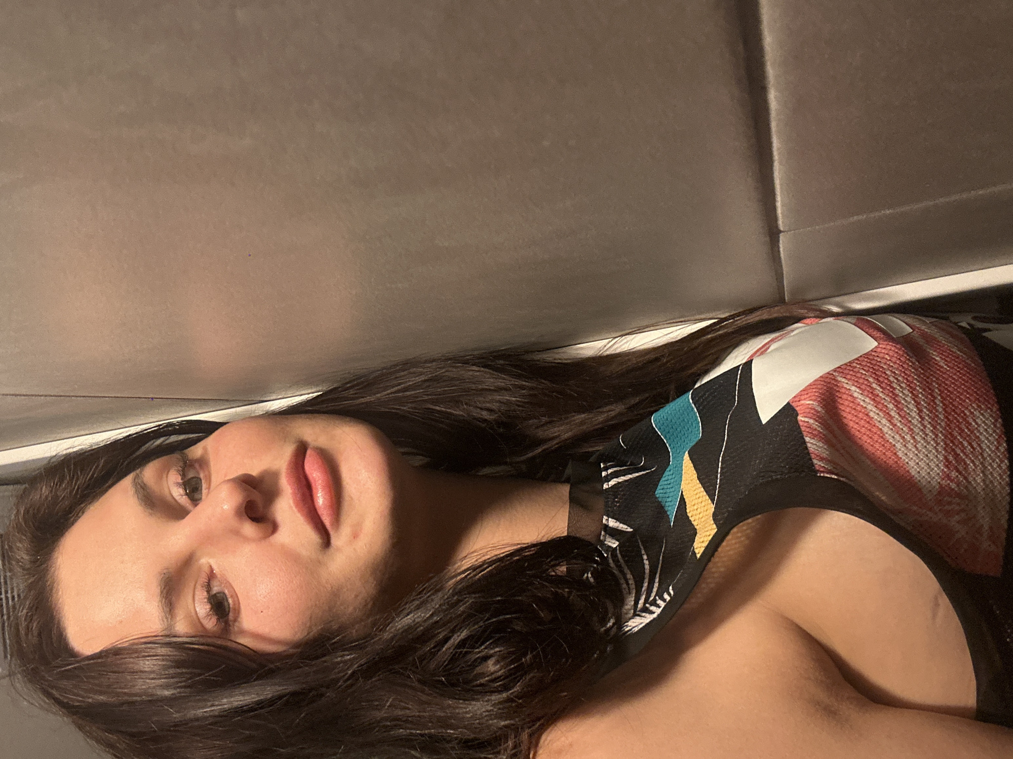 A young adult woman with dark brown hair and fair skin is captured in a close-up, angled shot. She is leaning against a textured gray wall, her head tilted slightly. She is wearing a black top with a colorful, abstract pattern featuring white, red, teal, and yellow elements. Her right shoulder and a portion of her chest are visible. The background is a plain, gray, padded surface with horizontal seams. The lighting is warm and somewhat dim, casting soft shadows.