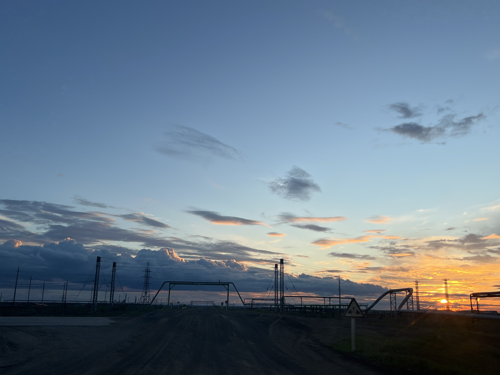 This image depicts an industrial landscape at sunset in Khar'yaginskii, Russia. The sky transitions from a clear blue at the top to hues of orange and yellow near the horizon, illuminated by the setting sun. Scattered clouds, tinged with pink and orange, are visible against the blue. In the foreground, a dirt road leads towards a complex of industrial structures. These include tall transmission towers, pipes supported by A-frame structures, and other unidentified industrial equipment. A yellow hazard sign is visible on the right side of the road. The overall impression is one of an industrial area at dusk, with the natural beauty of the sunset contrasting with the man-made structures.