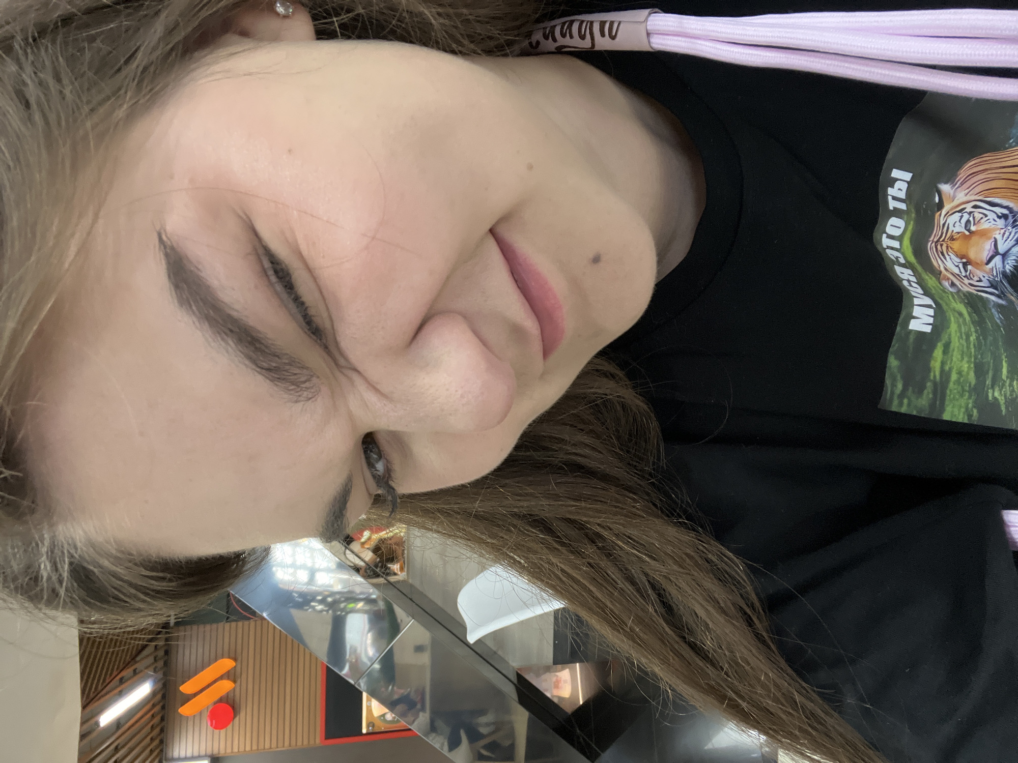A young woman with long brown hair is taking a selfie. She is wearing a black t-shirt with a tiger graphic and Russian text that reads "Муся это ты" (Musya, is that you?). She is winking and scrunching her face in a playful expression. In the background, an orange and red "M" logo is visible, suggesting the location is a McDonald's restaurant in Yekaterinburg, Russia.