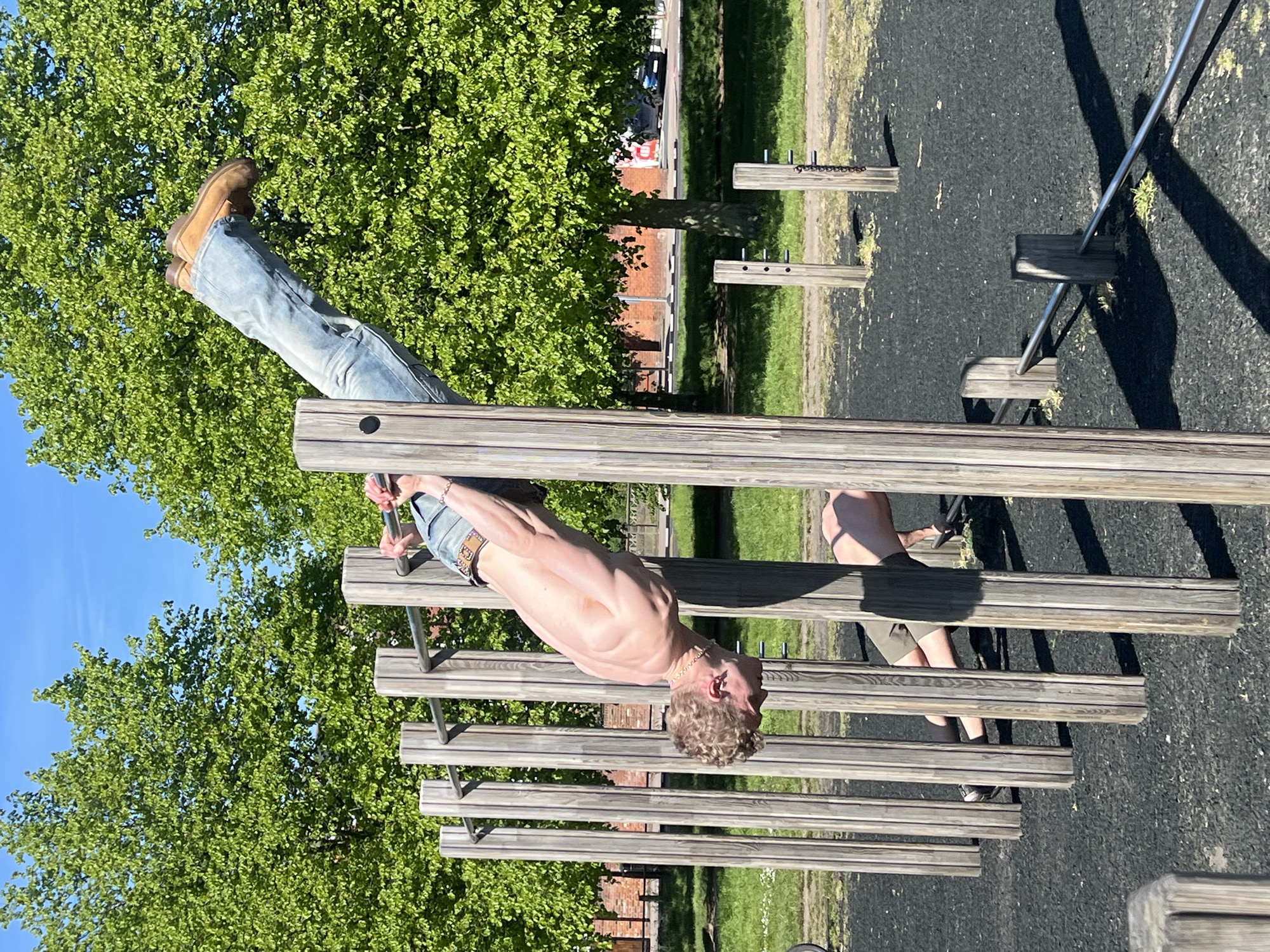 A man hangs upside down on a set of wooden parallel bars in an outdoor setting, possibly a park or playground. He is shirtless, wearing jeans and brown boots. His body is oriented vertically, with his head pointing towards the ground. His arms are extended, gripping the bars. Another individual with shorts is visible on the ground. The background includes green trees, a brick building, and a play area with black safety flooring. The scene appears to be set in daylight under a clear sky. The location might be a public park in London, United Kingdom.