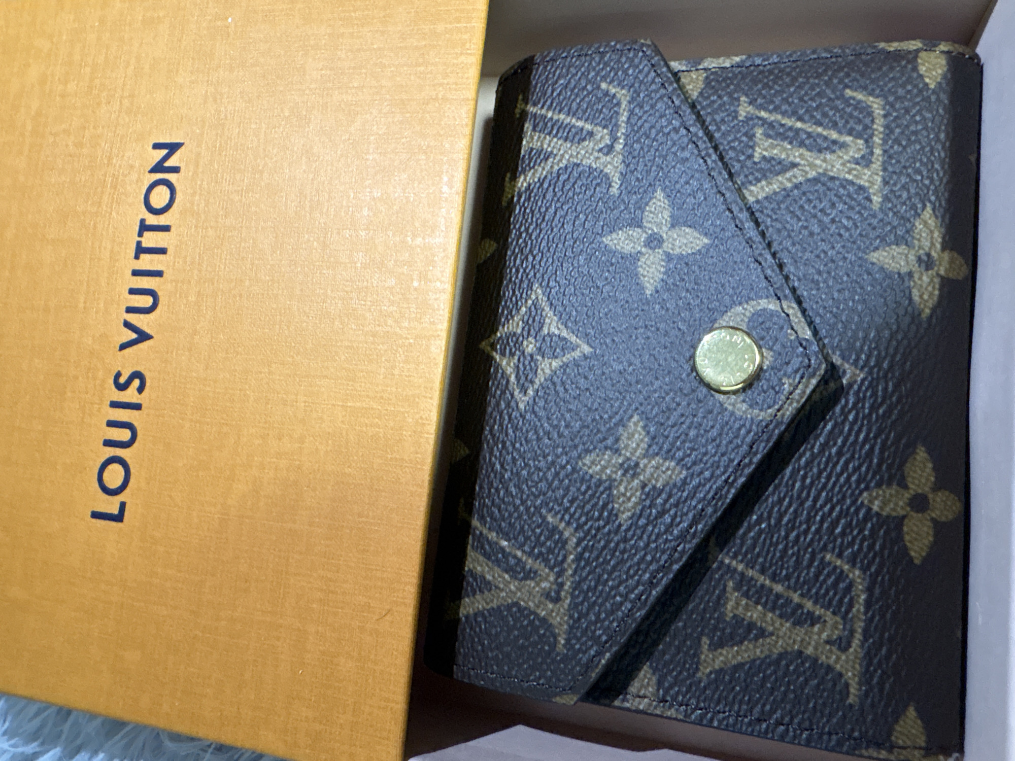 The image shows a Louis Vuitton wallet inside its box. The box is tan with the words "LOUIS VUITTON" printed vertically in dark blue. The wallet has a brown monogram pattern and a gold-colored snap closure. The wallet and box are positioned inside a white inner box. The scene provides no information on a location in Cheshnegirovo, Bulgaria.