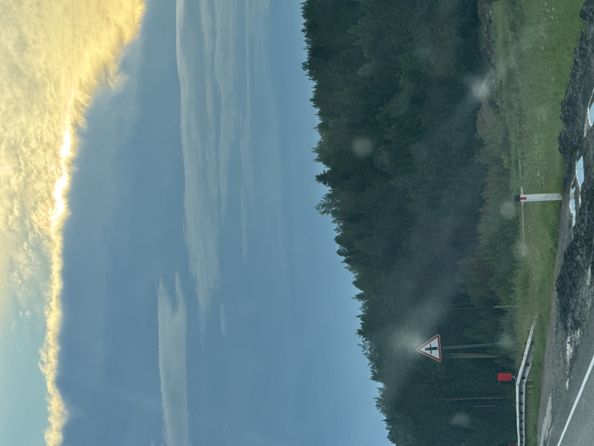 The image depicts a roadside scene in Karagay, Russia, under a cloudy sky with streaks of yellow sunlight illuminating the upper atmosphere. The foreground shows a paved road with white lane markings, bordered by a guardrail and grassy verges. Several small, light-colored puddles are visible on the road surface. A triangular road sign with a red border and a black cross symbol on a white background is prominently displayed on a post. To the left of the sign, a red rectangular object is partially visible. In the background, a dense forest of dark green coniferous trees stretches across the horizon, with a lighter, hazy layer of foliage closer to the road. The overall environment suggests a rural or semi-rural setting, likely during the daytime.