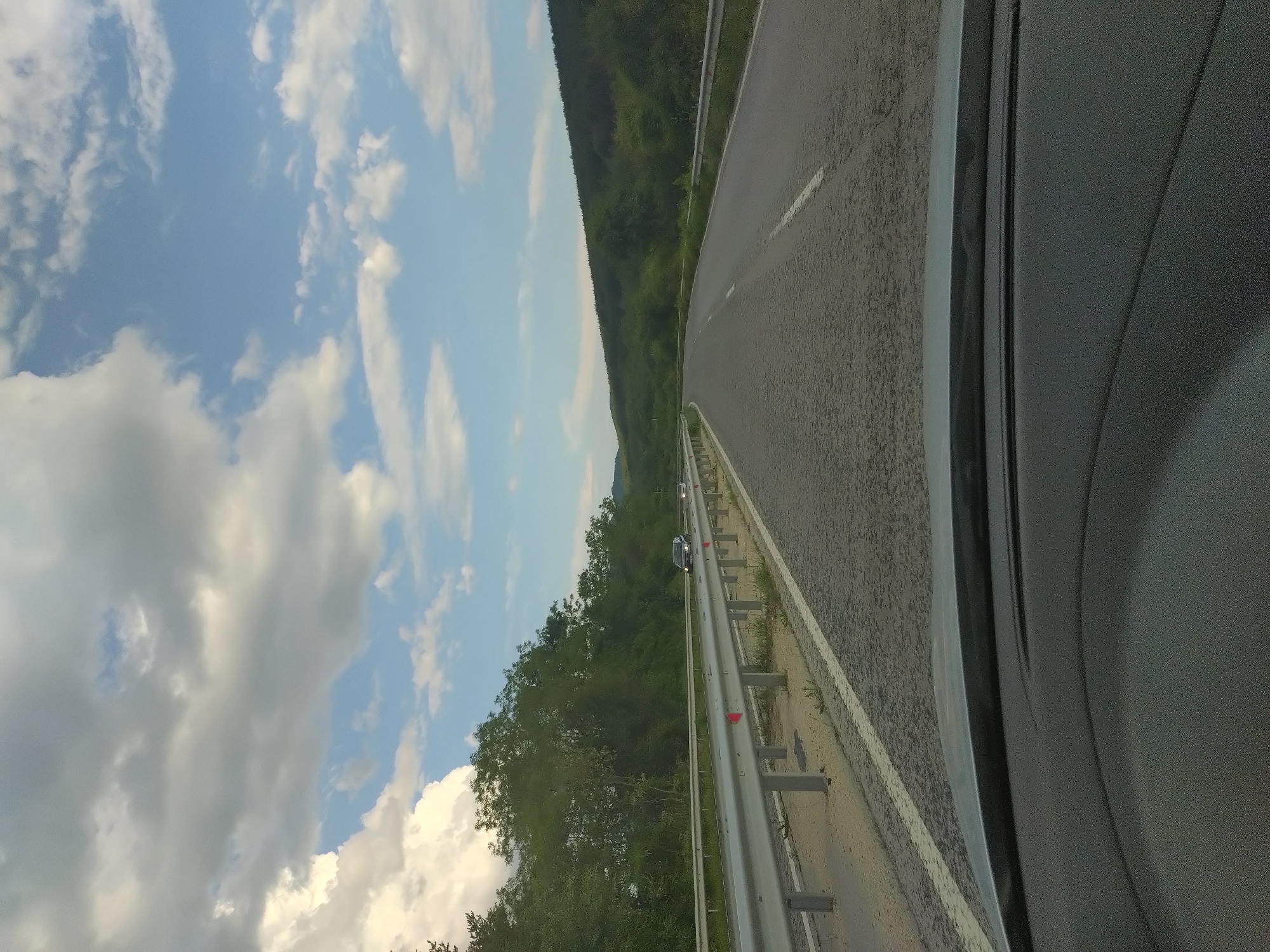 The image depicts a forensic view from the interior of a moving vehicle, specifically from the driver's or front passenger's perspective, looking forward over a dark, textured dashboard and the upper rim of a steering wheel. The vehicle is traversing a two-lane asphalt road in the geographical area of Zmeyovo, Bulgaria. The road surface, which shows a coarse texture in the foreground, features a continuous white line delineating the right shoulder and a dashed white line separating the two lanes. The road appears to curve moderately to the right in the middle ground.
A galvanized steel guardrail, equipped with regularly spaced red reflective markers, parallels the left side of the road. Beyond this barrier lies a strip of unpaved terrain with minimal vegetation, followed by a dense expanse of green trees, forming a forest. Two distinct passenger vehicles are observable in the opposing lane, approaching the viewer: a dark-colored sedan in the mid-ground and a lighter-colored vehicle positioned further behind it.
On the right side of the road, an identical galvanized steel guardrail extends along the shoulder. Immediately beyond this barrier, a natural incline rises, densely covered with green foliage, forming a heavily forested hillside.
The sky above displays a combination of cloud cover and clear blue patches. Large, bright white cumulus clouds are predominantly visible in the upper left quadrant, while smaller, more diffused white clouds are scattered across the remaining sky, revealing discernible sections of unclouded blue. The illumination suggests natural daylight conditions. No human figures are present within the captured scene.