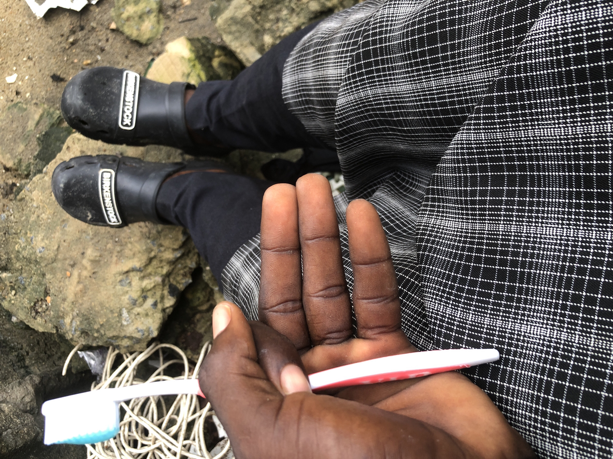 A medium shot from a high angle shows a person's hand holding a white and red toothbrush with a blue head. The hand is dark-skinned and positioned in the center-left of the frame. The toothbrush is resting on a black and white checkered fabric. The person is wearing black pants and black clogs with "BIRKENSTOCK" visible on the sides. The background consists of rocks and some coiled white wire, suggesting an outdoor or possibly a construction environment. The location is identified as Ajah, Nigeria.