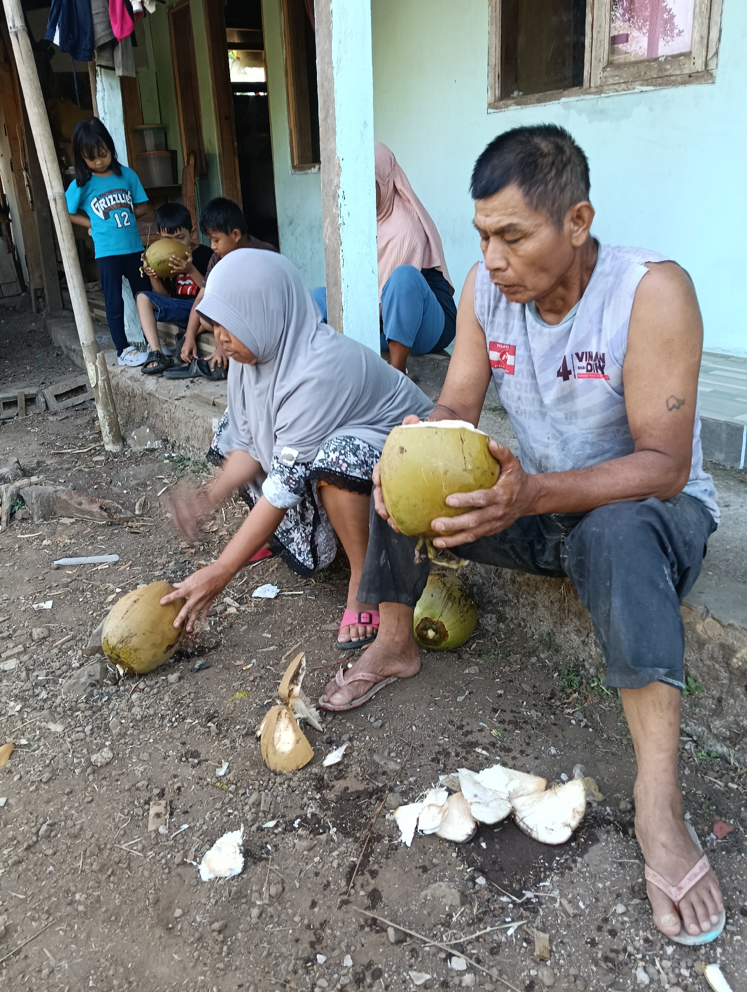In Tasikmalaya, Indonesia, an outdoor scene unfolds with a group of people preparing coconuts. A man, seated on a step, holds a partially opened coconut in his hands, his gaze directed downwards. Beside him, a woman in a hijab also sits, her hands positioned over another coconut on the ground, suggesting she is in the process of opening it. Scattered around them are pieces of coconut husk and flesh, along with two whole coconuts, one of which is cut open. To the left, three children are visible. One child, wearing a blue t-shirt with "GRIZZLIES 12" printed on it, stands looking down, while two others are seated, one holding a coconut. The environment appears to be a simple outdoor area with a dirt ground, a concrete step, and parts of a building with light blue walls and wooden window frames in the background. Clothes hang on a bamboo pole to the left. The overall impression is one of domestic activity and community.
