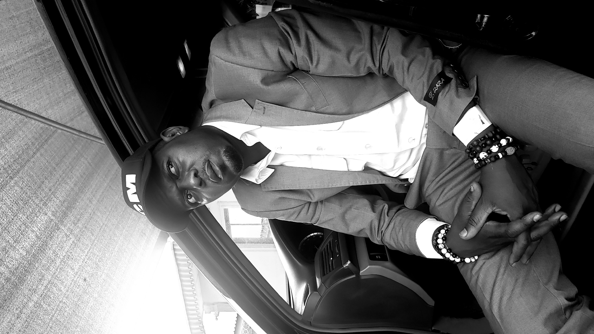A monochromatic image captures a man dressed in a suit, sitting inside a car in Sabon Tasha, Nigeria. The image is oriented sideways, with the man looking directly towards the viewer. He wears a light-colored suit jacket over a white collared shirt and matching trousers. A black baseball cap, featuring a prominent white "M" logo, is worn on his head. His expression is calm, with a slightly upward gaze.
Both of the man's wrists are adorned with multiple beaded bracelets. These accessories include a mix of black, white, and silver beads, some of which feature patterned or decorative charms, like a silver-toned bead with a swirl design. On the left cuff of his suit jacket, the brand name "ZARA" is clearly visible in a distinct font. The background elements reveal parts of the car's interior, including sections of the dashboard and a door panel. Bright light streams in from outside the vehicle, suggesting the photo was taken during daylight hours. The man's hands are casually clasped together in his lap.