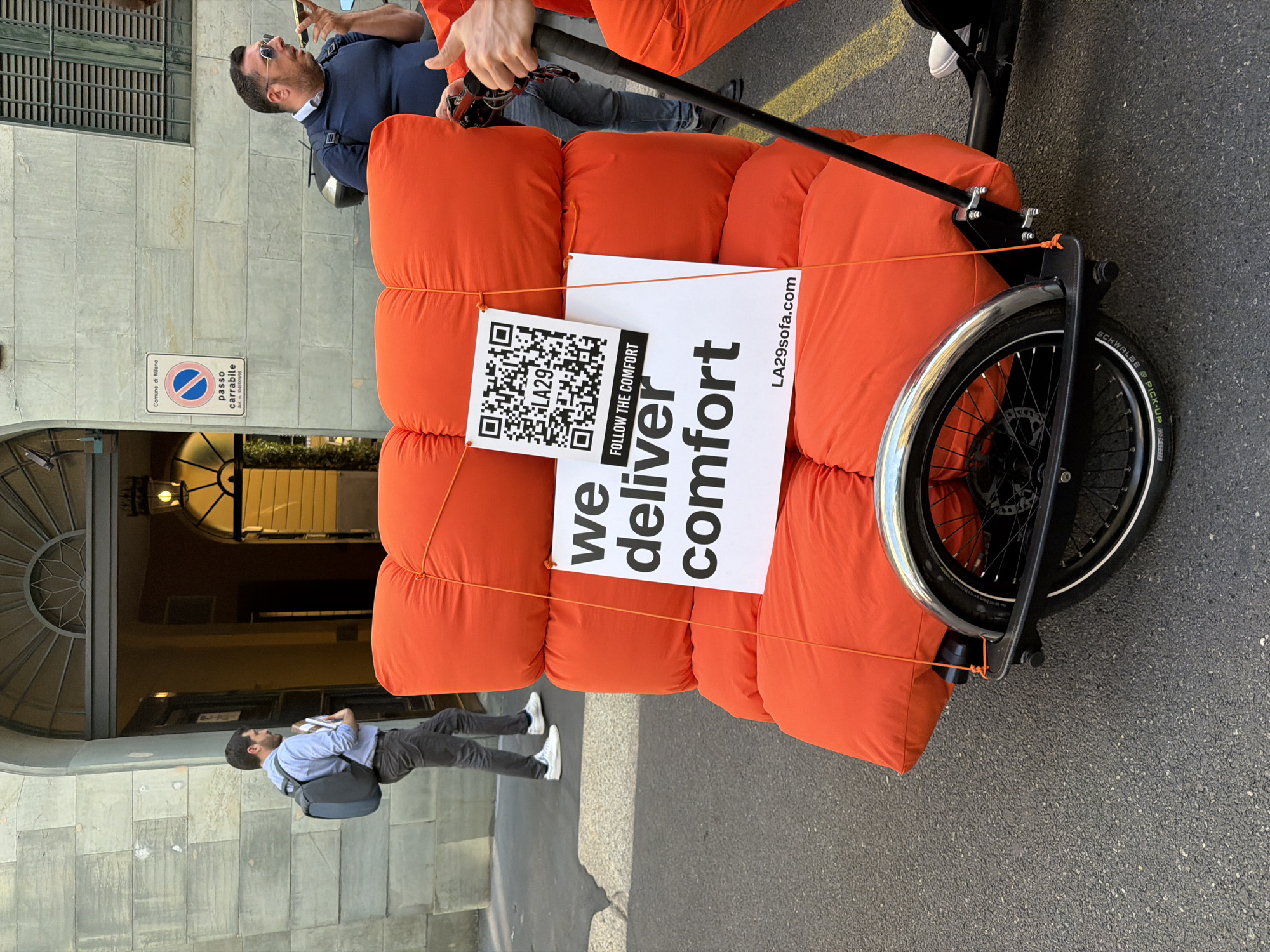 The image depicts an urban street scene in Milan, Italy, during bright daylight. The central subject is a large, vibrant orange, modular sofa or collection of cushions, being transported on a custom-built, single-wheeled trolley. The sofa segments are plush and appear to be tied together with orange cords, secured to the trolley's frame.
A white rectangular sign is prominently displayed on the sofa, featuring large text that reads "we deliver comfort." Above this, smaller text states "FOLLOW THE COMFORT," and a QR code with "LA29SOFA" next to it is visible. The website "LA29sofa.com" is also printed on the sign.
A person, partially visible on the right, wears orange pants matching the sofa and a dark top, holding the long handle of the trolley, suggesting they are moving it. The trolley has a black frame, a single front wheel with a silver fender, and appears robust.
In the background, traditional Italian architecture with stone-like facades and an arched entryway frames the left side of the scene. A "Comune di Milano" sign is mounted on a building, clearly displaying "passo carrabile" (driveway/no parking). Two other individuals are visible: one man in a light blue shirt and backpack walks away in the mid-ground, while another man in a dark polo shirt and sunglasses looks upwards in the further background. The ground is paved with asphalt. The event appears to be a promotional activity or an unconventional delivery.