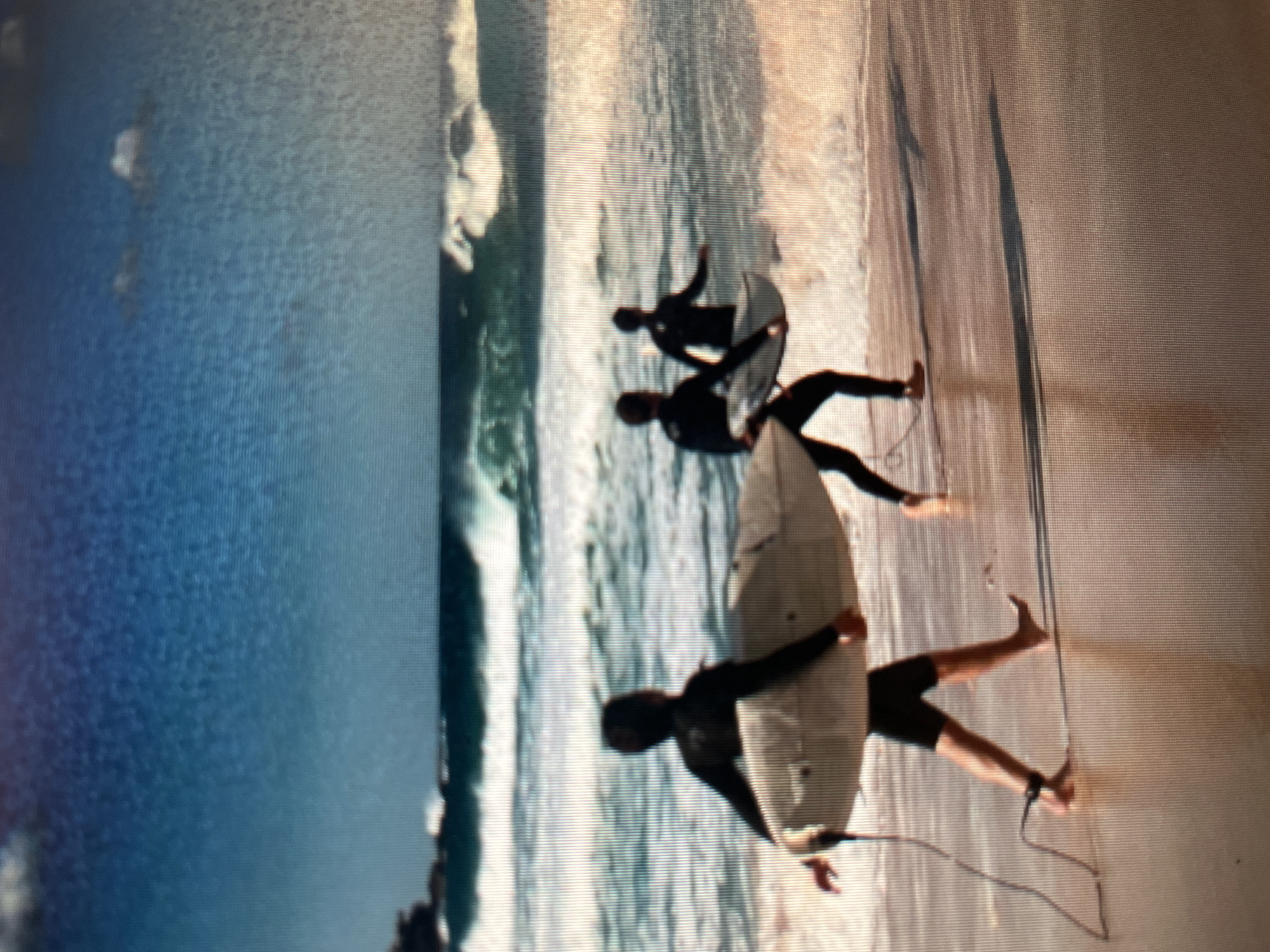 Three surfers in black wetsuits walk along a sandy beach toward the ocean. The surfer in the foreground is carrying a white surfboard horizontally. The waves are breaking in the background, and the sky is blue with some clouds. The scene appears to be taking place in Sejahtera, Indonesia, a coastal city known for its surf breaks.