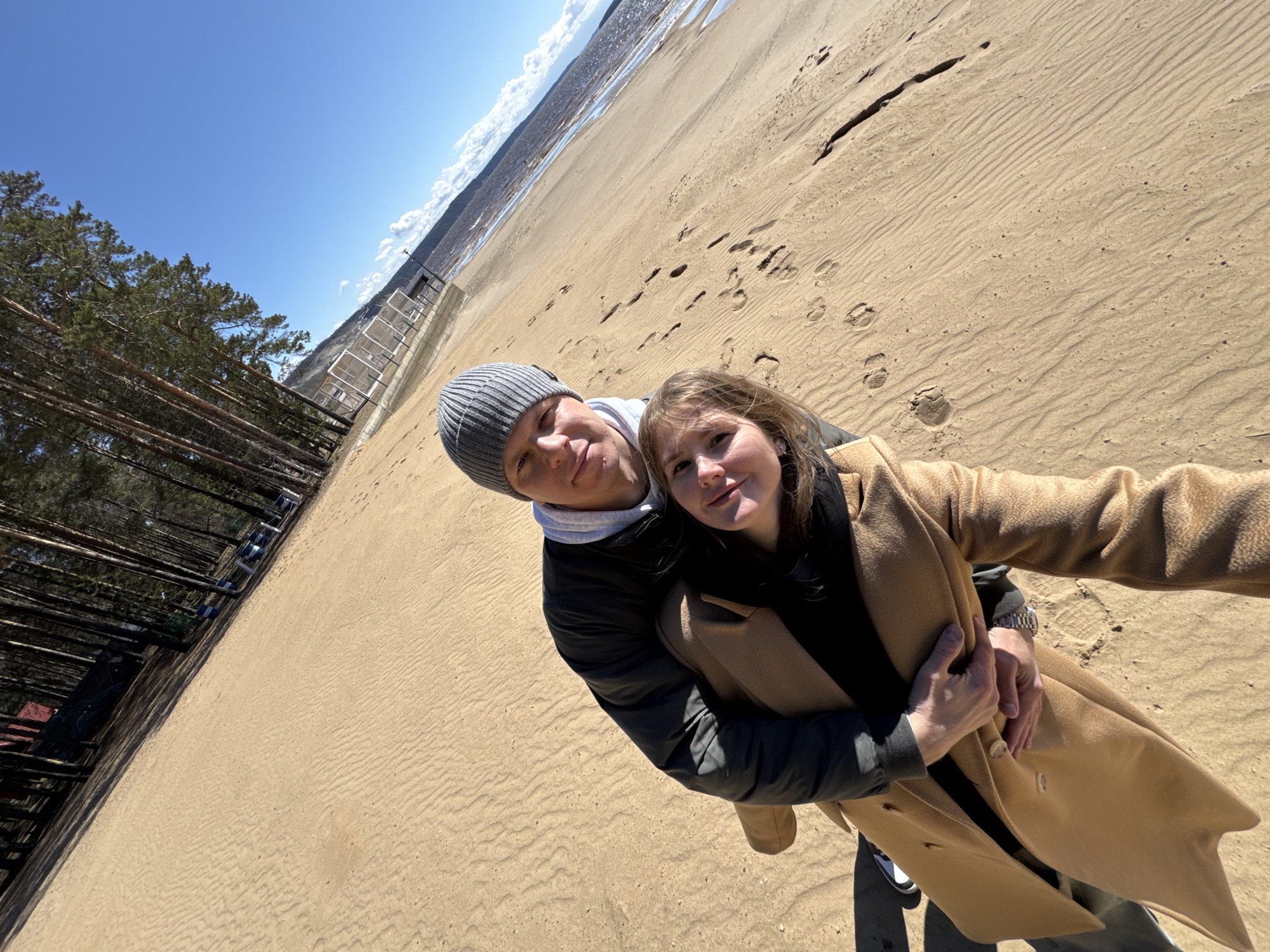 This selfie-style image captures a young couple on a wide, sandy beach under a bright blue sky. The man, on the left, wears a grey beanie, a light grey hoodie, and a dark jacket, while embracing the woman. The woman, on the right, sports a camel-colored long coat over a dark top. Both individuals are smiling gently at the camera, conveying a sense of happiness and affection.
The setting features a vast expanse of sandy ground with clear ripple patterns and numerous footprints, suggesting recent activity. To the left, a dense line of tall, slender pine trees provides a natural backdrop. In the mid-ground, a large body of water, likely the Kama Reservoir given the location context of Polazna, Russia, is visible, reflecting the bright sky. Distant structures, possibly fences or boardwalk elements, line the far edge of the sandy shore. The sky is clear with a few scattered white clouds. The bright daylight indicates it is likely mid-morning or afternoon. Despite the sunny conditions, the couple's warm clothing suggests cool weather. The photo has a distinctive tilted perspective, typical of a wide-angle selfie, encompassing both the subjects and their expansive surroundings. There is no visible text in the image.