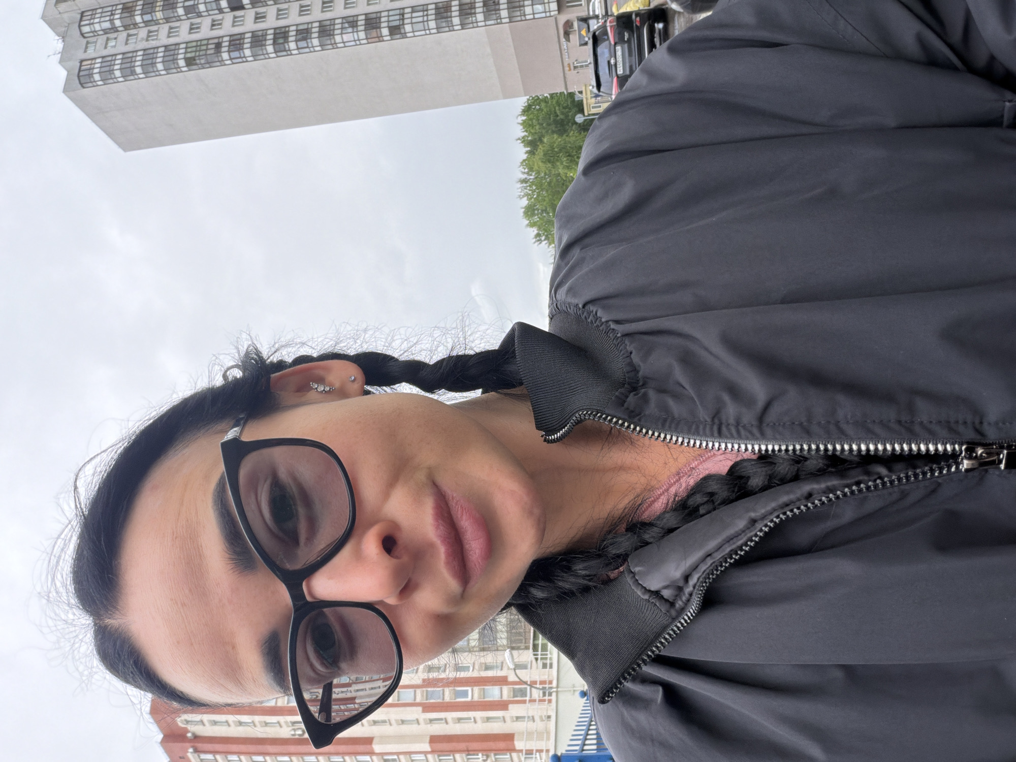 A selfie shot taken outdoors in what appears to be Saint Petersburg, Russia. The main subject is a person wearing black-rimmed glasses and a dark bomber jacket zipped to mid-chest, with black braids. A pink top is visible underneath the jacket. A tall gray building with numerous windows is visible behind and to the left of the person, and a red and white building can be seen behind and to the right. The sky is overcast. Cars are parked nearby and visible in the background. The photo is rotated slightly counter-clockwise.