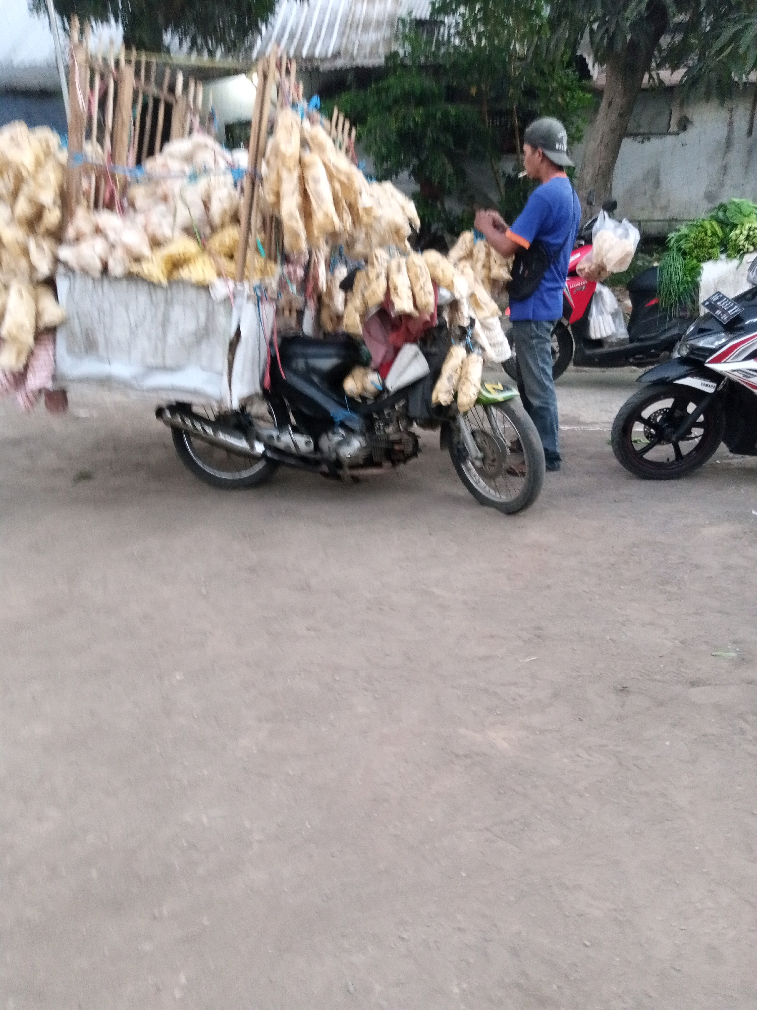 A medium-shot, slightly blurry, horizontal outdoor photograph captures a street vendor selling goods from a motorcycle in Sono Ageng, Indonesia. The vendor, a man wearing a blue t-shirt, dark pants, and a grey baseball cap, stands with his back slightly to the viewer, holding a cigarette in his mouth and looking down at the goods. He has a dark sling bag across his chest. The motorcycle, a black and red scooter with a license plate beginning with "AG", is loaded with numerous bags of what appear to be dried snacks or food items, attached to a wooden frame erected over the vehicle. The goods are a light tan color and are bundled or bagged. In the background, another motorcycle is parked to the right, and a pile of green vegetables can be seen next to it. A wooden fence or structure is visible behind the vendor and his goods. The ground is a dirt or gravel surface. The overall scene depicts a typical street vending setup in a rural or semi-urban Indonesian setting.