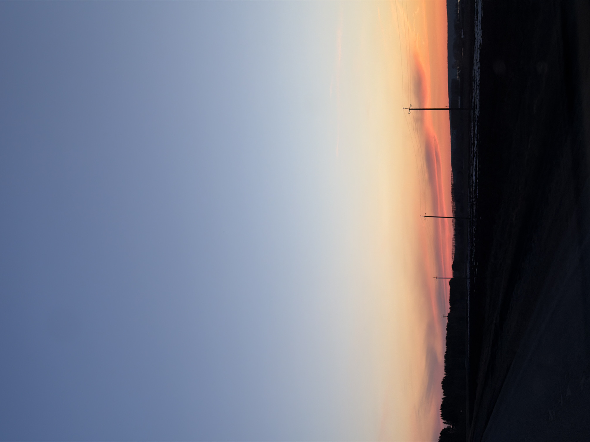 The image shows a landscape at dusk or dawn, likely in a rural area, although a specific location in Eldushino, Russia, cannot be determined without further contextual information. The sky transitions from a deep blue on the left side of the frame to a gradient of orange, pink, and peach colors on the right, indicating a sunrise or sunset. Several utility poles are silhouetted against the colorful sky. A dark strip along the bottom edge of the frame represents the ground, possibly a field or road, while a darker forested or built-up area lies above. No people, actions, or interactions are visible.
