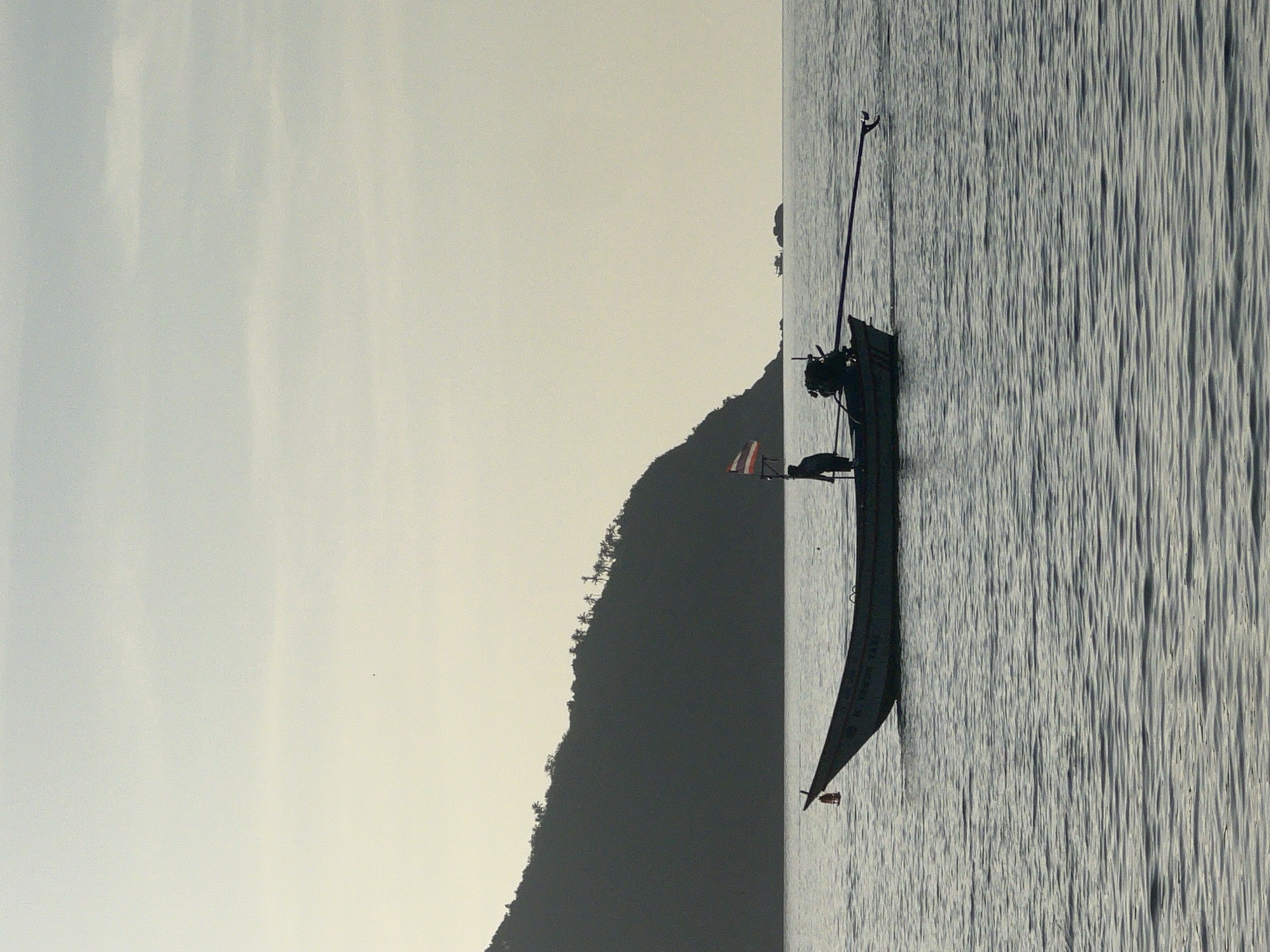 A single, silhouetted longtail boat with a person standing in it travels on calm water. The boat has a Thai flag, an engine with a long shaft extending to the right, and a small red lantern hanging from the front. In the background, a large, dark, tree-covered island dominates the left side of the frame, sloping down to the water. The sky is a pale, hazy yellow, with no visible clouds. The scene appears to be a coastal area in Ko Pha-ngan District, Thailand, likely at dawn or dusk.