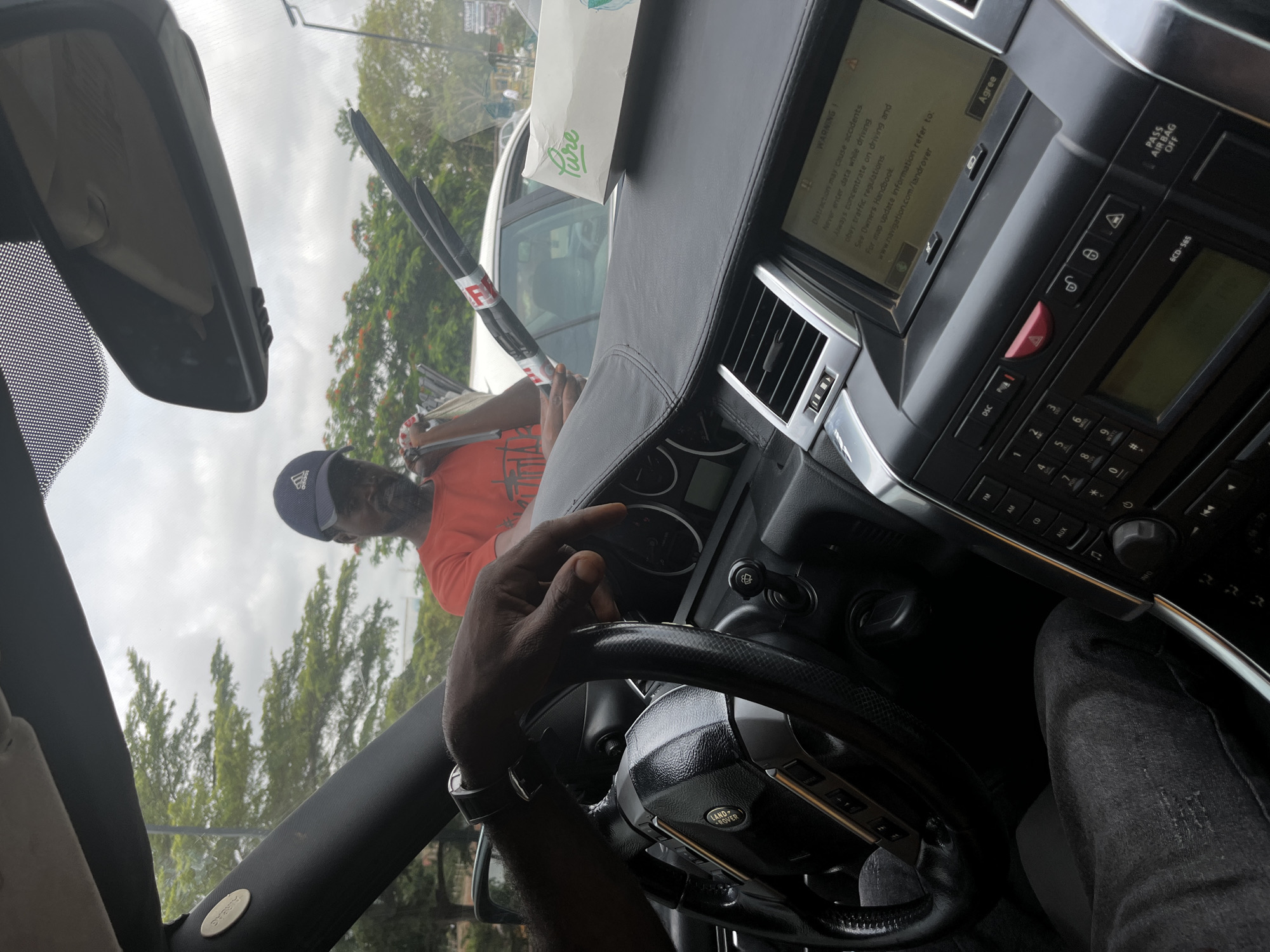This media file captures a street interaction from inside a car in Abuja, Nigeria, during the daytime on an overcast day.
The primary subject is the interior of a black Land Rover vehicle, featuring a dark-skinned person's hands on the "LAND ROVER" branded steering wheel. The driver is wearing a watch on their left wrist and dark, possibly distressed, jeans. The dashboard is visible, topped with a box of "Pure" brand tissues. The car's infotainment screen displays a safety "WARNING!" message: "Distractions may cause accidents. Never enter data while driving. Always concentrate on driving and obey traffic regulations. See Owners Handbook. www.navigation.com/landrover. For map update information refer to: Agree". Below the screen, the center console features various buttons, including "DSC", "AUX", a red hazard light button, a numeric keypad, and text such as "6CD-565" and "PASS AIR BAG OFF".
Outside the car's windshield, another dark-skinned person is partially visible, wearing an orange shirt and a blue "Adidas" baseball cap. This individual appears to be engaging with the car's occupant, holding what looks like a squeegee or long brush and possibly some magazines or newspapers. Part of a white car is visible in the background, along with green trees and reddish-orange flowering foliage under an overcast sky. The activity suggests a common street scene where vendors or service providers approach vehicles stopped at a light or in traffic.