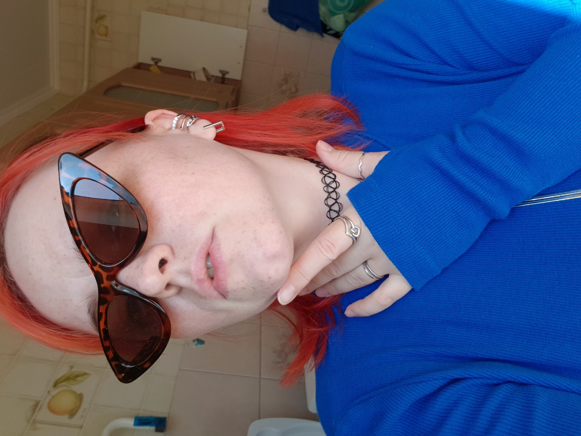 A person with red hair and wearing brown sunglasses and a blue ribbed sweater is indoors. The person has multiple piercings in their left ear and is wearing a black choker. The environment includes a bathroom with a tiled floor, a wooden cabinet, and a white toilet. The location is Unknown, Unknown.