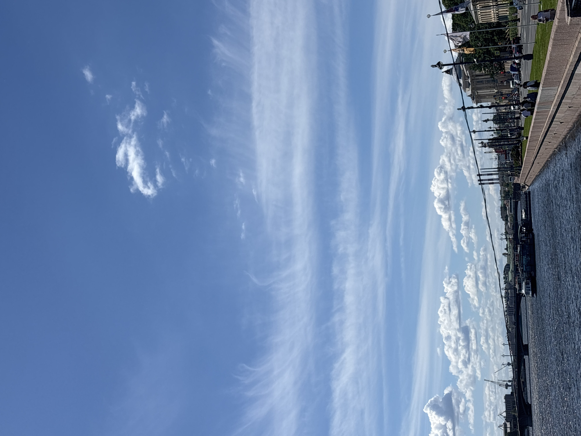 The image shows a waterfront view of the Neva River in Saint Petersburg, Russia, under a bright blue sky with wispy white clouds. On the left, a wide bridge spans the river, with cranes visible in the distance, suggesting a port area. A tour boat is docked on the right side of the river. The embankment on the right is lined with lampposts, flags, and pedestrians walking along a sidewalk. Beyond the sidewalk, a street with traffic and ornate buildings can be seen. The water of the Neva River is a deep blue with ripples. The scene appears to be a sunny day in a bustling urban environment.