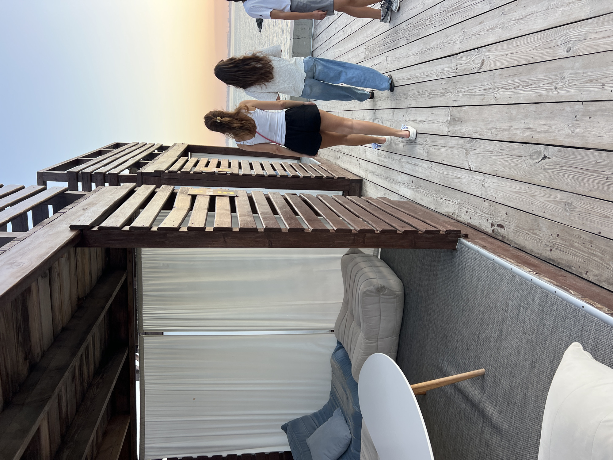 The image depicts an outdoor seating area with a wooden deck and a pergola structure. Inside the pergola, there are two upholstered seating elements, one resembling a futon and another a pouf, both in neutral tones. A white, oval-shaped coffee table with wooden legs is positioned in front of them. The area is partially enclosed by white curtains.
Outside the pergola, three individuals are walking on the wooden deck towards a body of water, likely the Black Sea, given the reference to Odesa, Ukraine. The sky displays a soft gradient of colors, suggesting either sunrise or sunset. The architecture of the pergola features horizontal wooden slats, creating a partial enclosure. The overall setting appears to be a relaxed, seaside establishment.