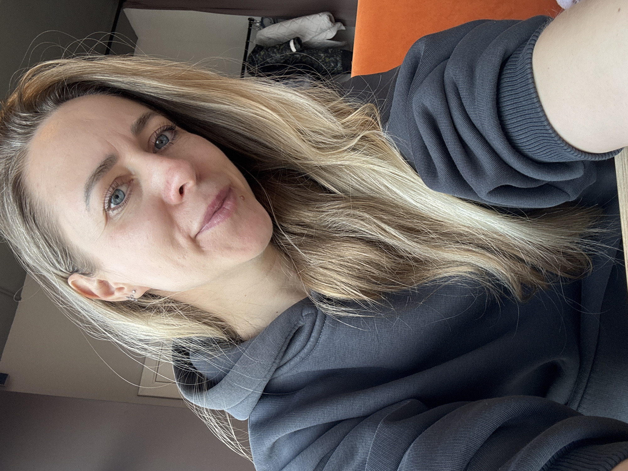 A woman with long blonde hair and blue eyes is shown in a selfie-style photo. She is wearing a dark gray hooded sweatshirt. The background is a room with an orange chair visible behind her. Other items are out of focus in the background. There is no specific information to identify the location as Chaikovsky, Russia.