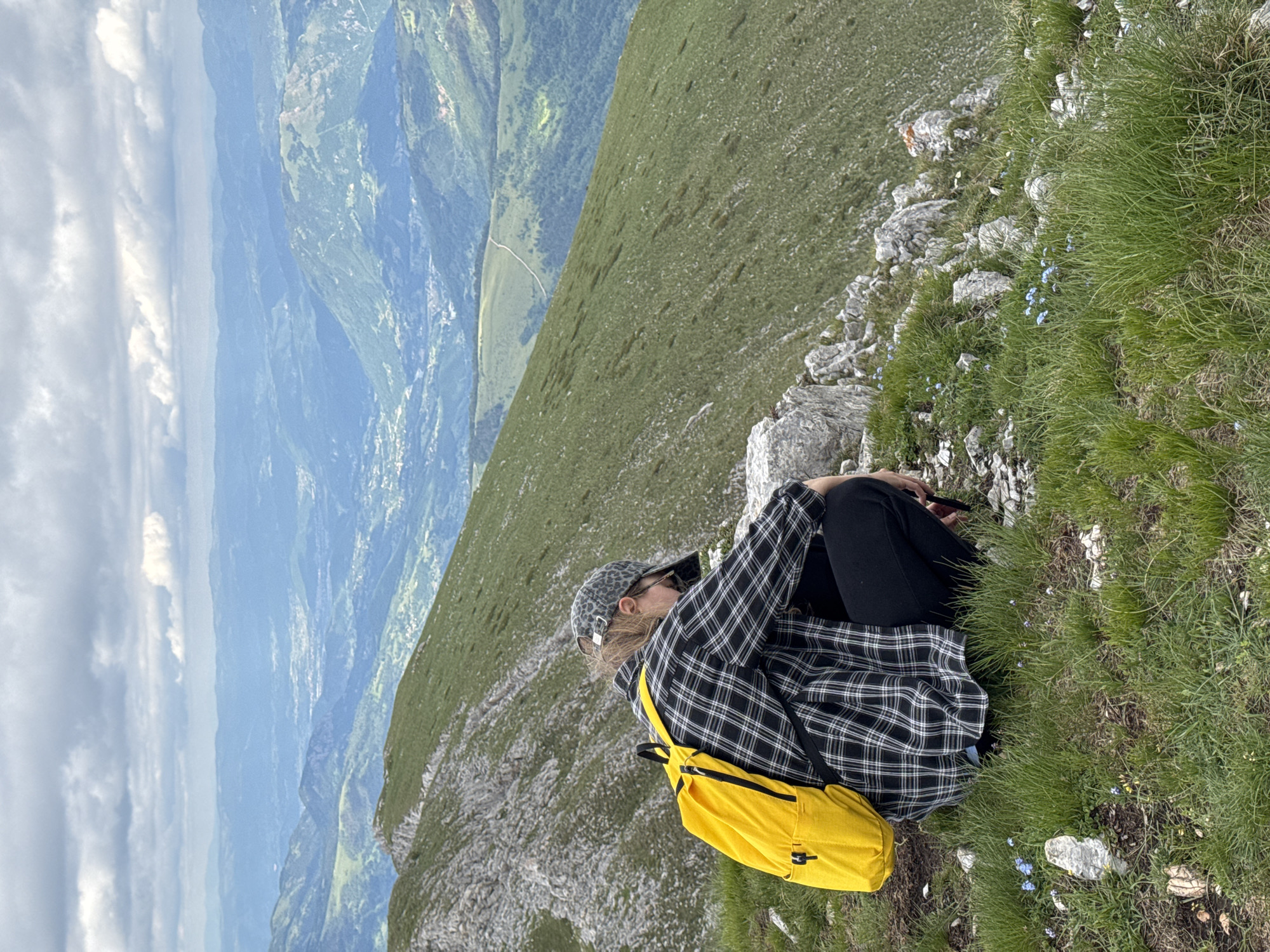 A person with a yellow backpack kneels on a grassy mountain slope, facing away from the camera. They wear a plaid shirt, black pants, and a leopard-print baseball cap. The background shows a vast mountainous landscape under a cloudy sky, with a valley containing a town or village in the distance. The location is the city of Cetinje, Montenegro.