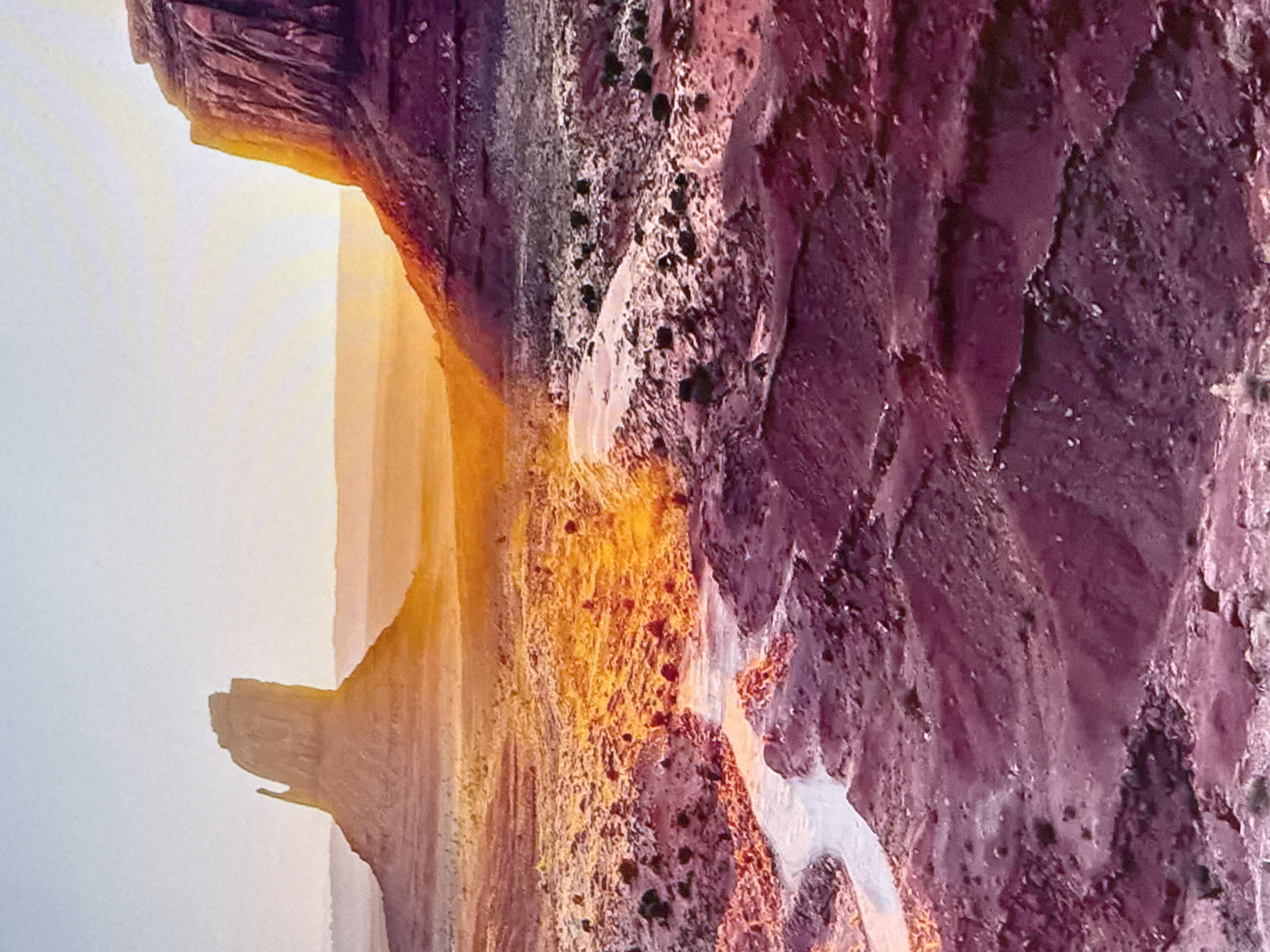 The image depicts a desert landscape at sunrise. Two large, distinctive rock formations dominate the scene. The one on the left is tall and narrow with a pointed top, while the one on the right is broader and more massive, with layered, reddish-brown rock faces. The sun is low on the horizon, casting a warm, golden glow across the landscape, particularly on the rock formations and the desert floor. A winding, sandy dry riverbed or wash snakes through the foreground and midground, reflecting the bright sunlight. The ground is a mix of reddish-brown earth, sparse vegetation, and scattered rocks. The sky is a pale blue, gradually fading to a lighter hue near the horizon. There are no people, animals, or man-made structures visible in the image. The scene is devoid of any urban elements. The location is not Simferopol, Ukraine, as the landscape is characteristic of the American Southwest, likely Monument Valley.