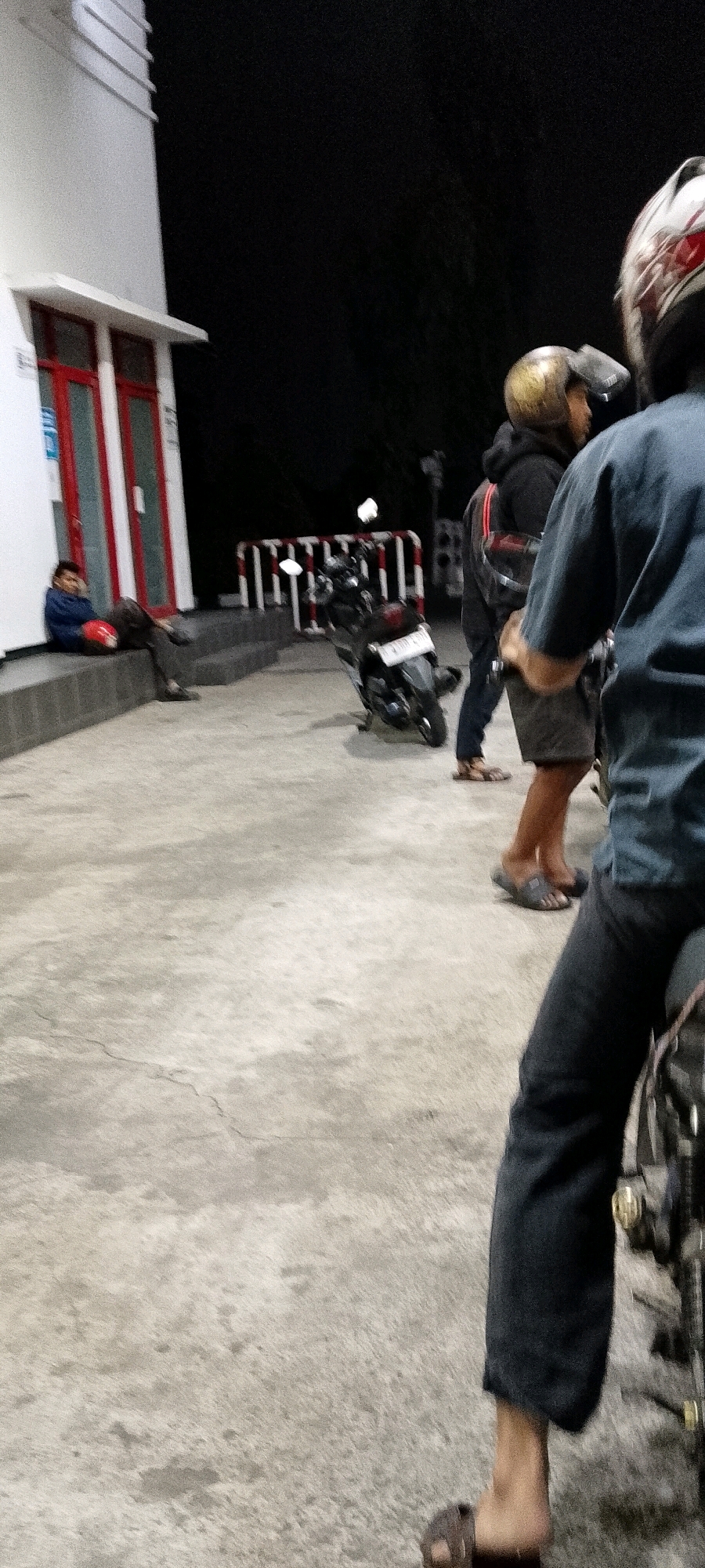 In Bekasi, Indonesia, at night, a person sits on a low concrete step in front of a white building with red-framed windows. The person is wearing a blue jacket and dark pants, with a red helmet resting on their lap. To the right, two people stand near a parked motorcycle. One person wears a gold helmet and a dark hoodie, while the other, seen from behind, is wearing a blue shirt and dark pants. The ground is a rough, light-colored concrete surface. A white and red striped barrier is visible in the background. The scene appears to be outdoors, possibly at a gas station or similar establishment given the architecture and lighting.