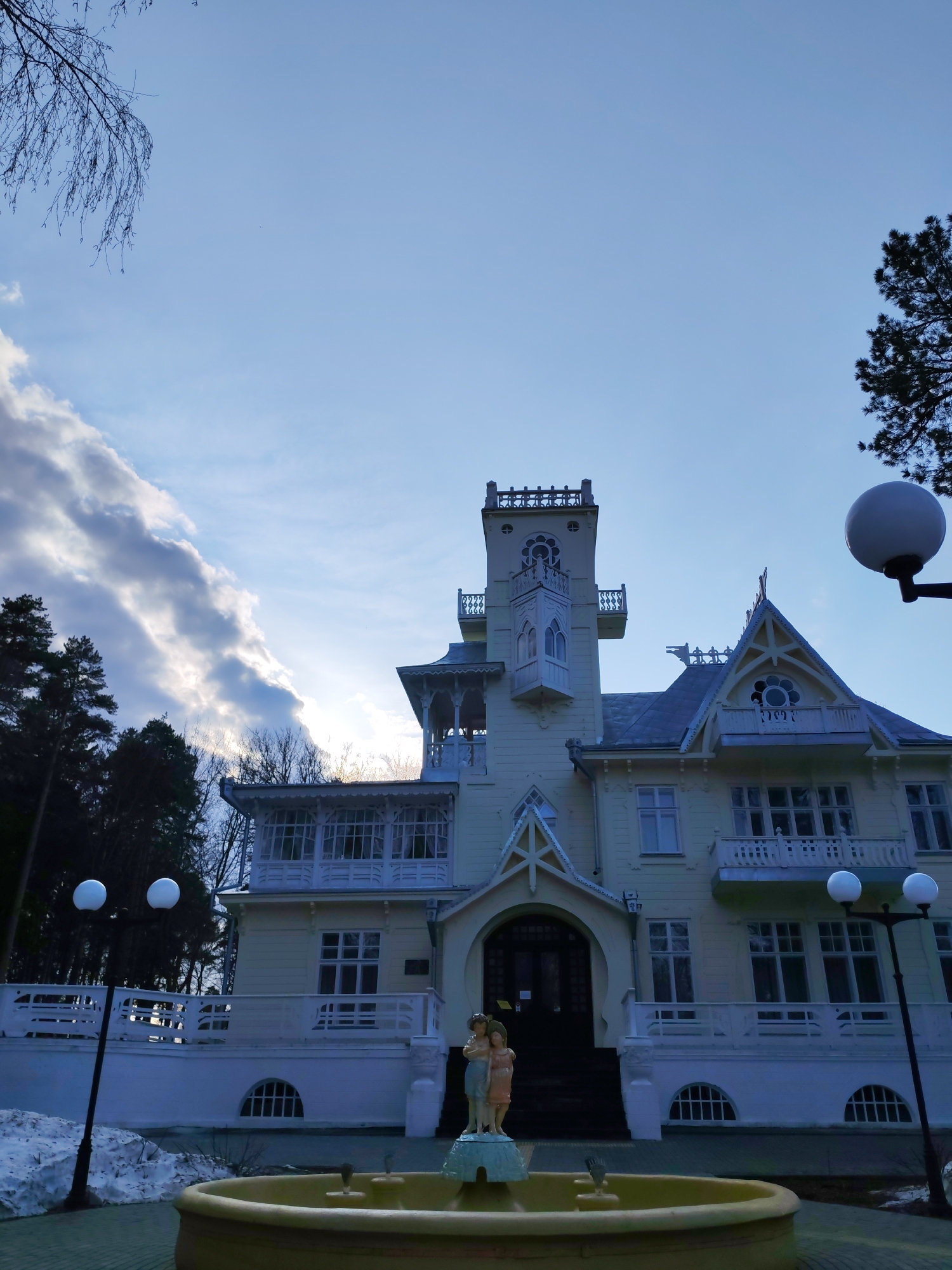 The image shows the Dacha Bashenina, an architectural landmark located in Sarapul, Russia. It is a light-colored, multi-story building with a prominent tower and ornate details. In front of the building is a fountain with a statue of two figures. The sky is partially cloudy. There are trees on either side of the building. Snow is visible on the ground to the left. Lamp posts with globe lights flank the path leading to the building. The photograph is taken from a low angle, looking up at the Dacha.